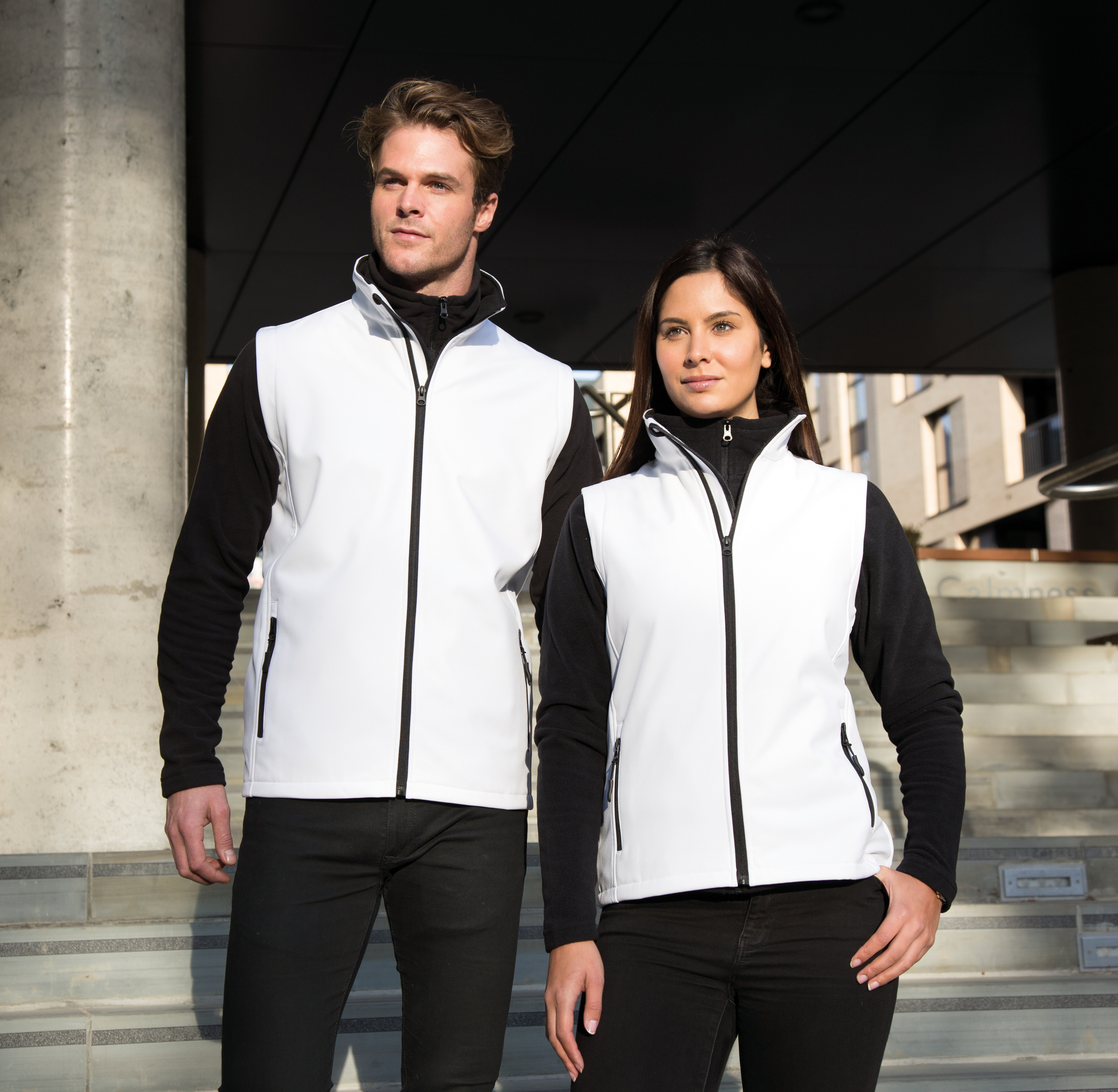 Hombre y mujer posando al aire libre con chalecos softshell blancos y capas negras sobre escaleras urbanas.