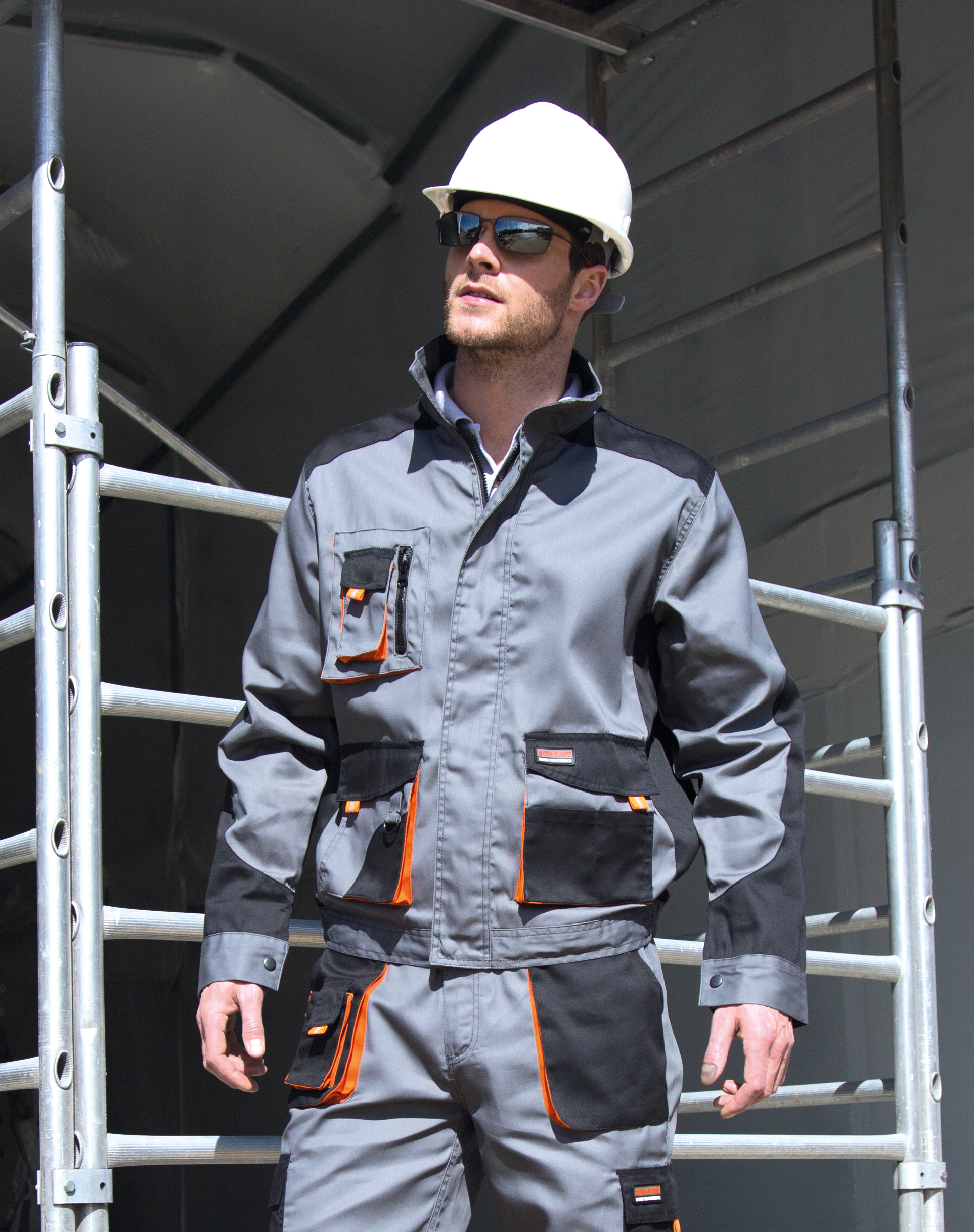 Trabajador con casco blanco y gafas de sol sobre un andamio vistiendo ropa de trabajo gris con bolsillos naranjas.