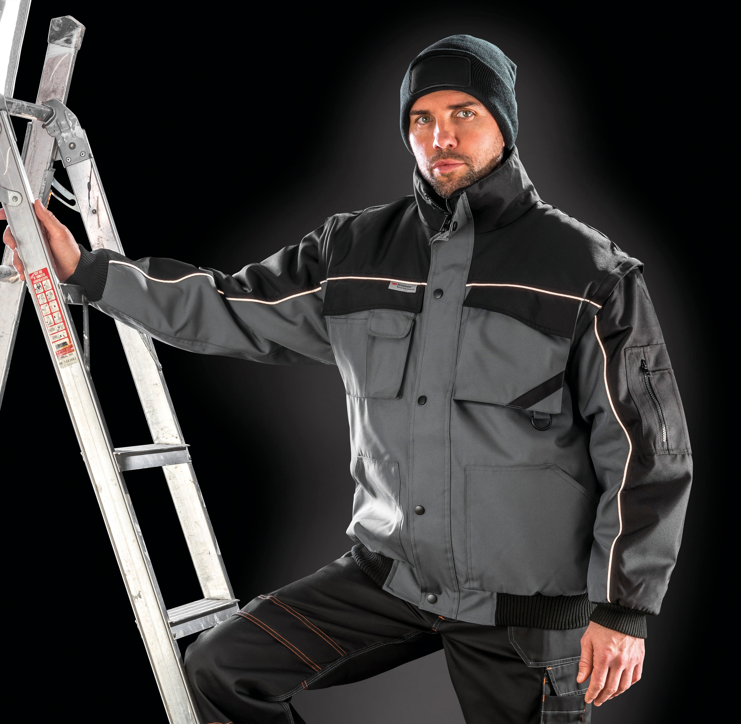 Hombre con chaqueta de trabajo acolchada y pantalones técnicos apoyado en una escalera metálica sobre fondo oscuro.