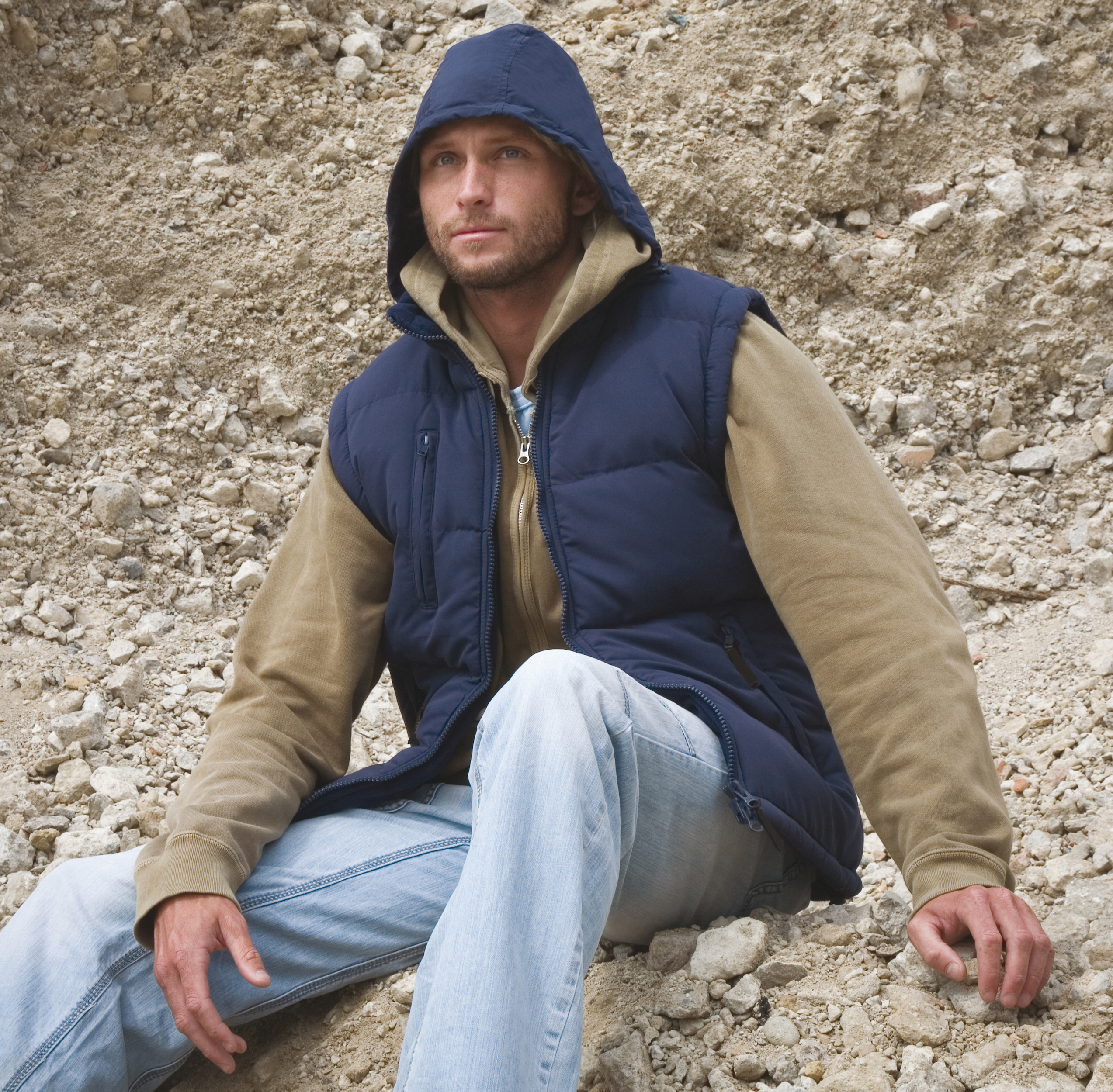 Hombre con chaleco acolchado y sudadera con capucha sentado sobre un terreno pedregoso