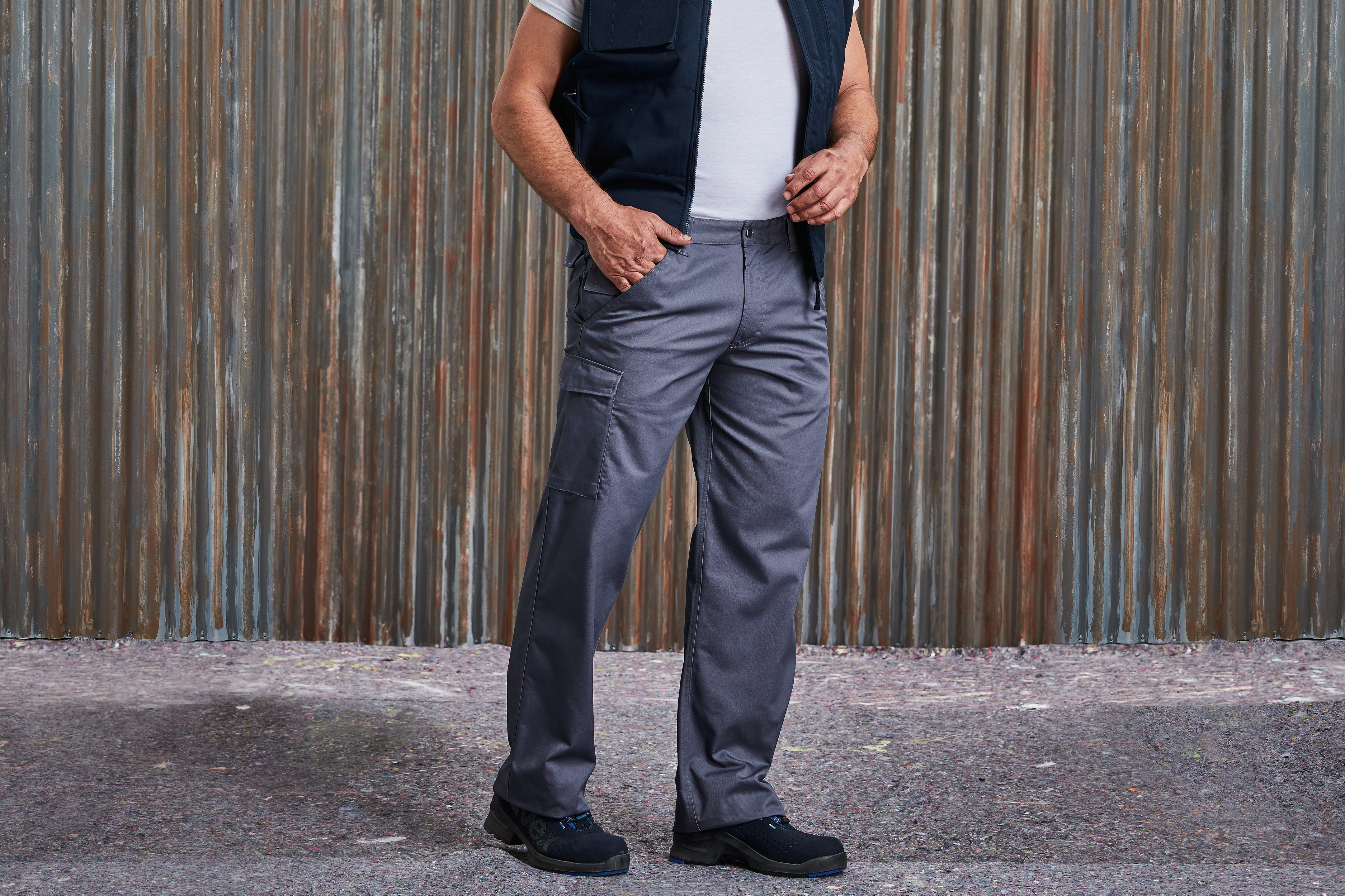 hombre con pantalones de trabajo grises con bolsillo lateral, chaleco azul marino, camiseta blanca y calzado de seguridad frente a un fondo de chapa metálica envejecida