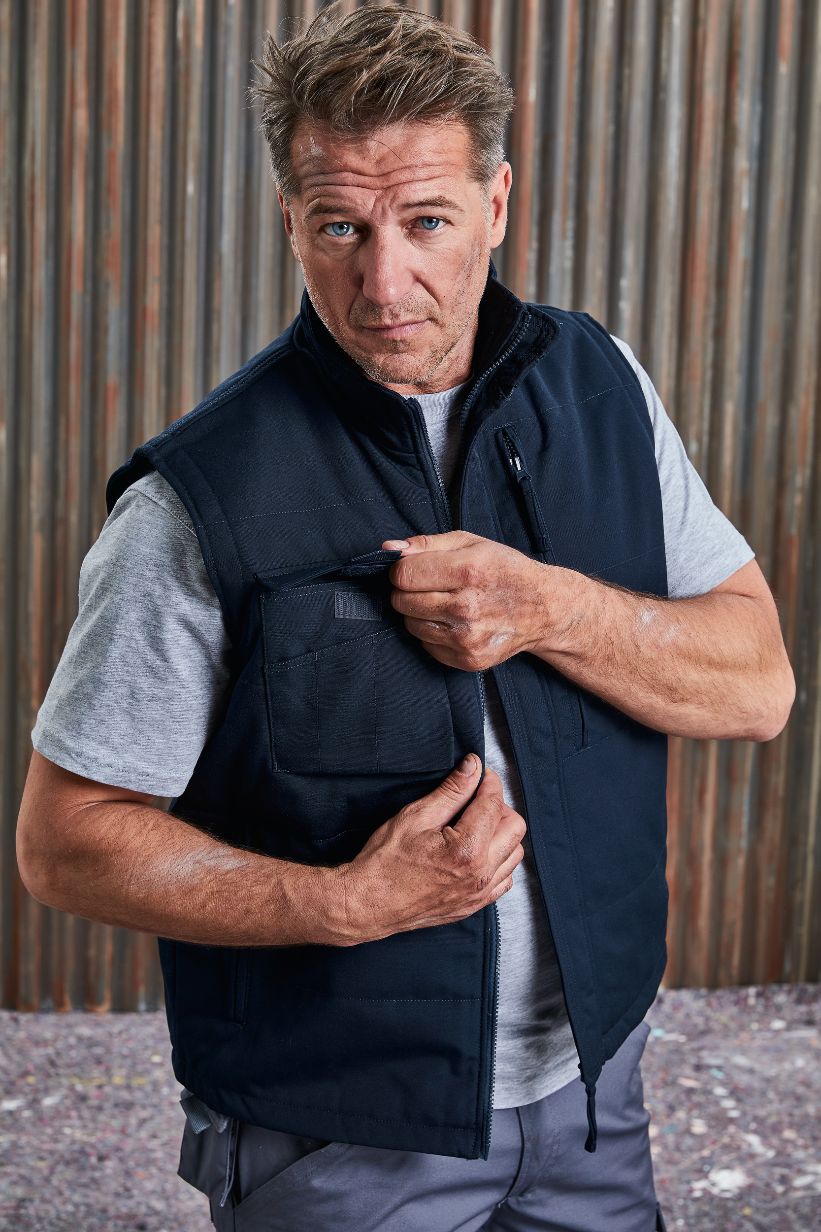 Hombre con chaleco de trabajo azul abriendo un bolsillo frontal frente a una pared metálica corrugada