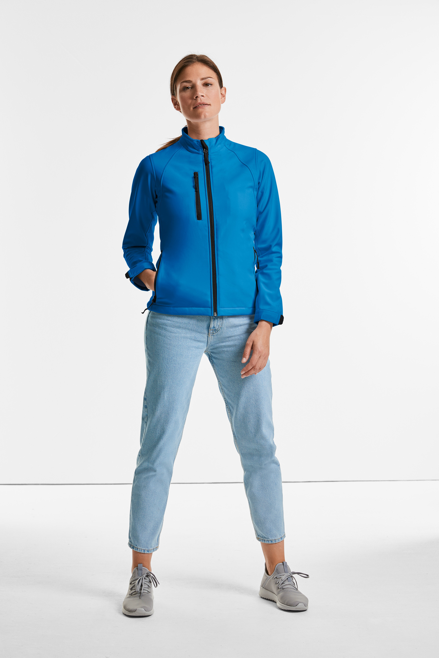 Mujer de pie con chaqueta softshell azul y vaqueros claros sobre fondo blanco