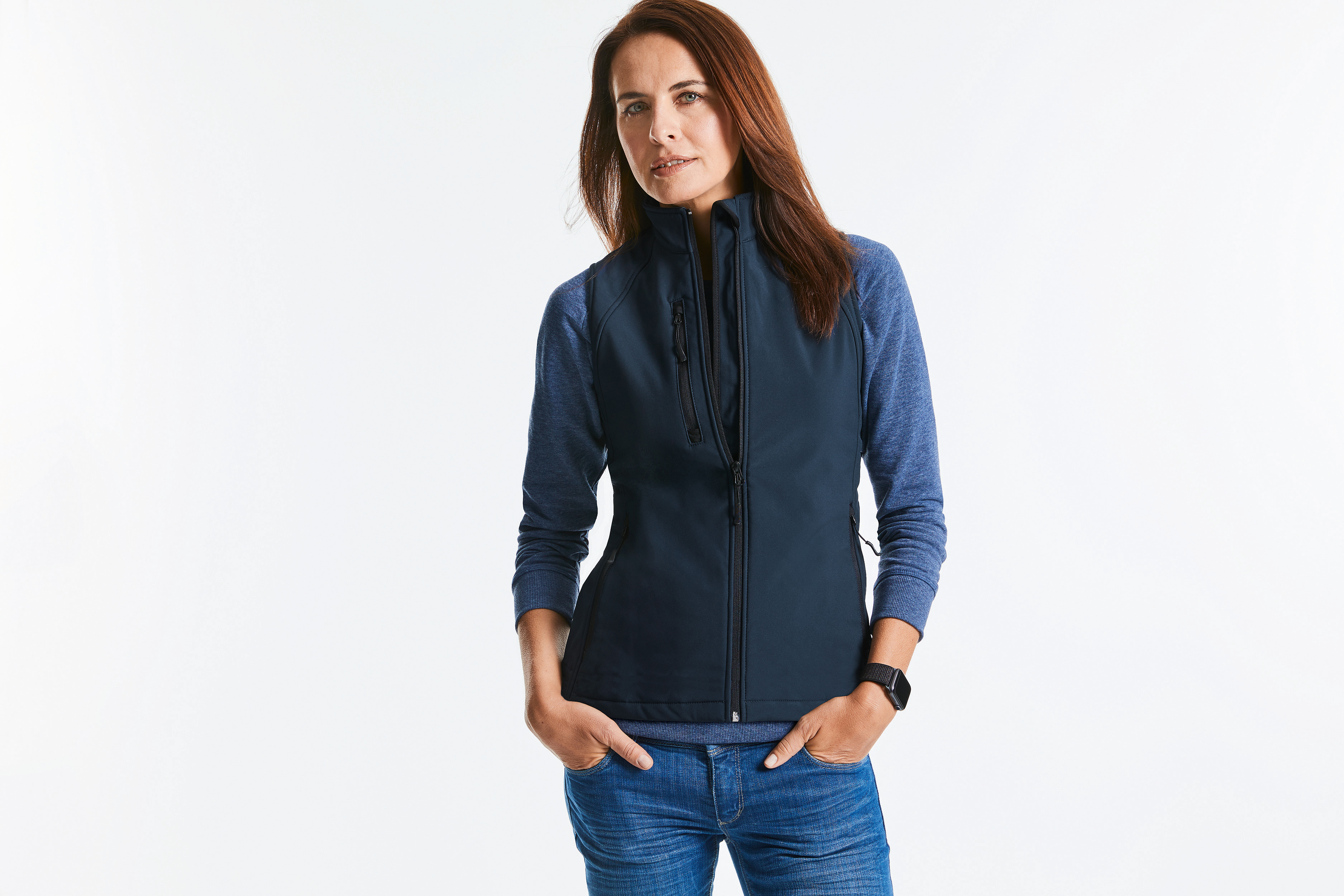 Mujer de pie con chaleco softshell azul marino, camiseta de manga larga y vaqueros sobre fondo blanco
