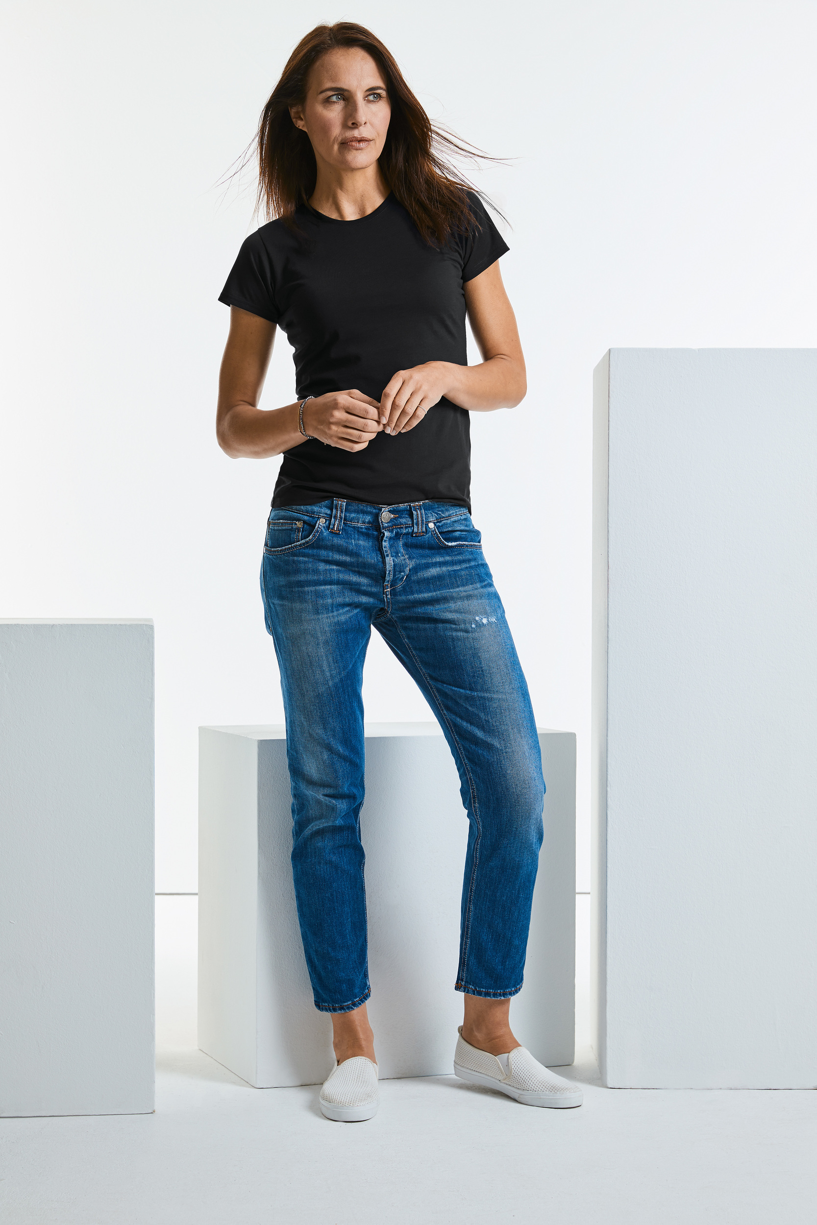 Mujer joven con camiseta negra, vaqueros azules y zapatillas blancas en un estudio minimalista con bloques blancos.