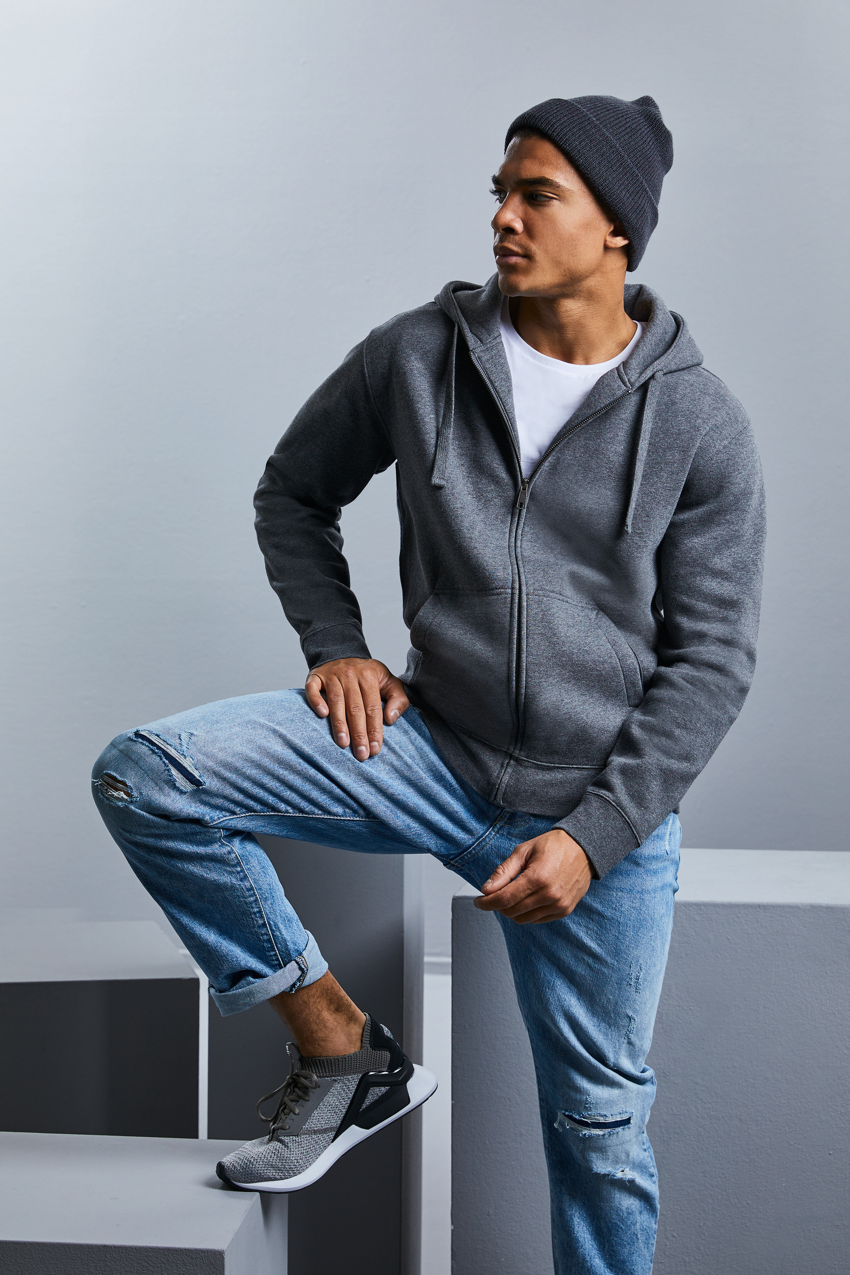 Hombre joven con sudadera gris con capucha, gorro beanie, vaqueros desgastados y zapatillas apoyando un pie en un bloque sobre fondo neutro.