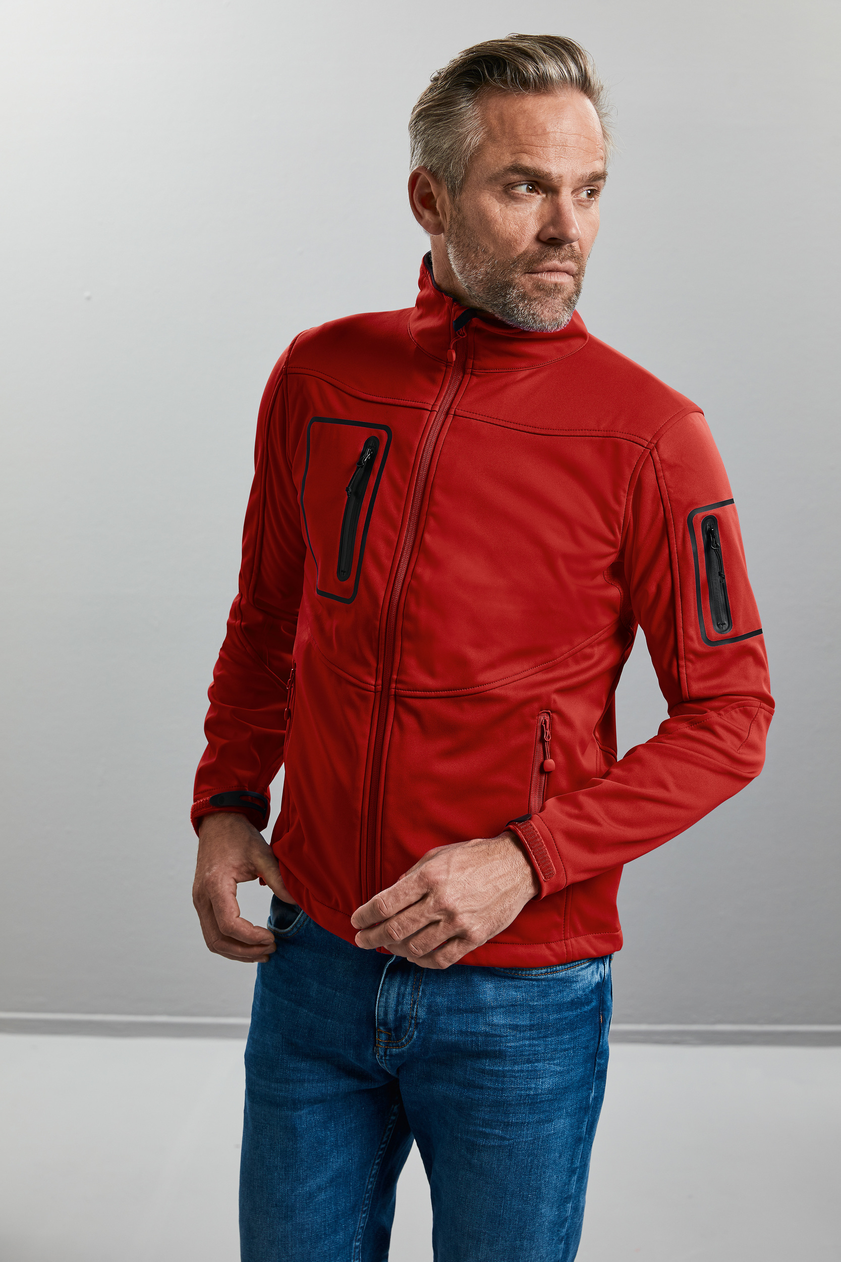 Hombre con chaqueta softshell roja y vaqueros en fondo neutro ajustándose la cremallera.