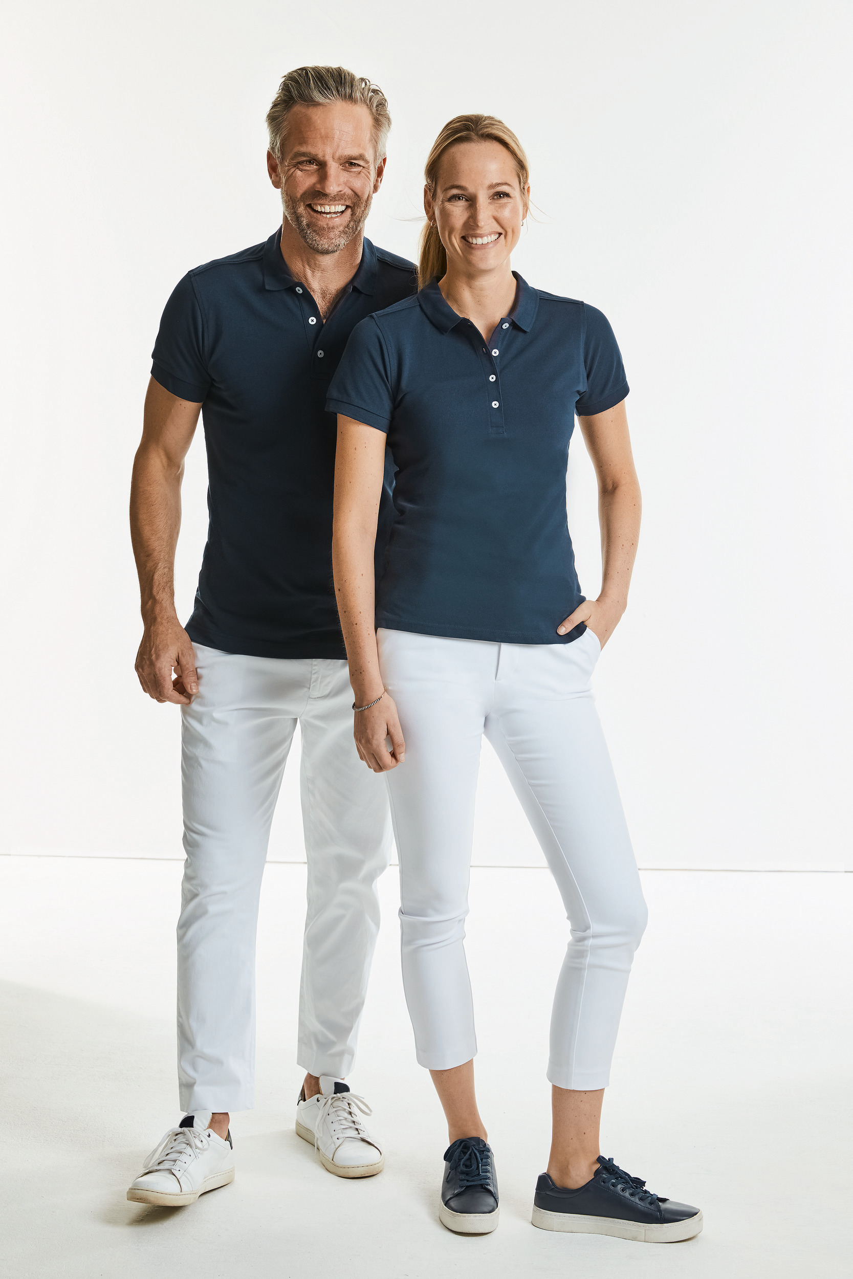 dos adultos sonrientes con polo azul marino y pantalones blancos sobre fondo blanco