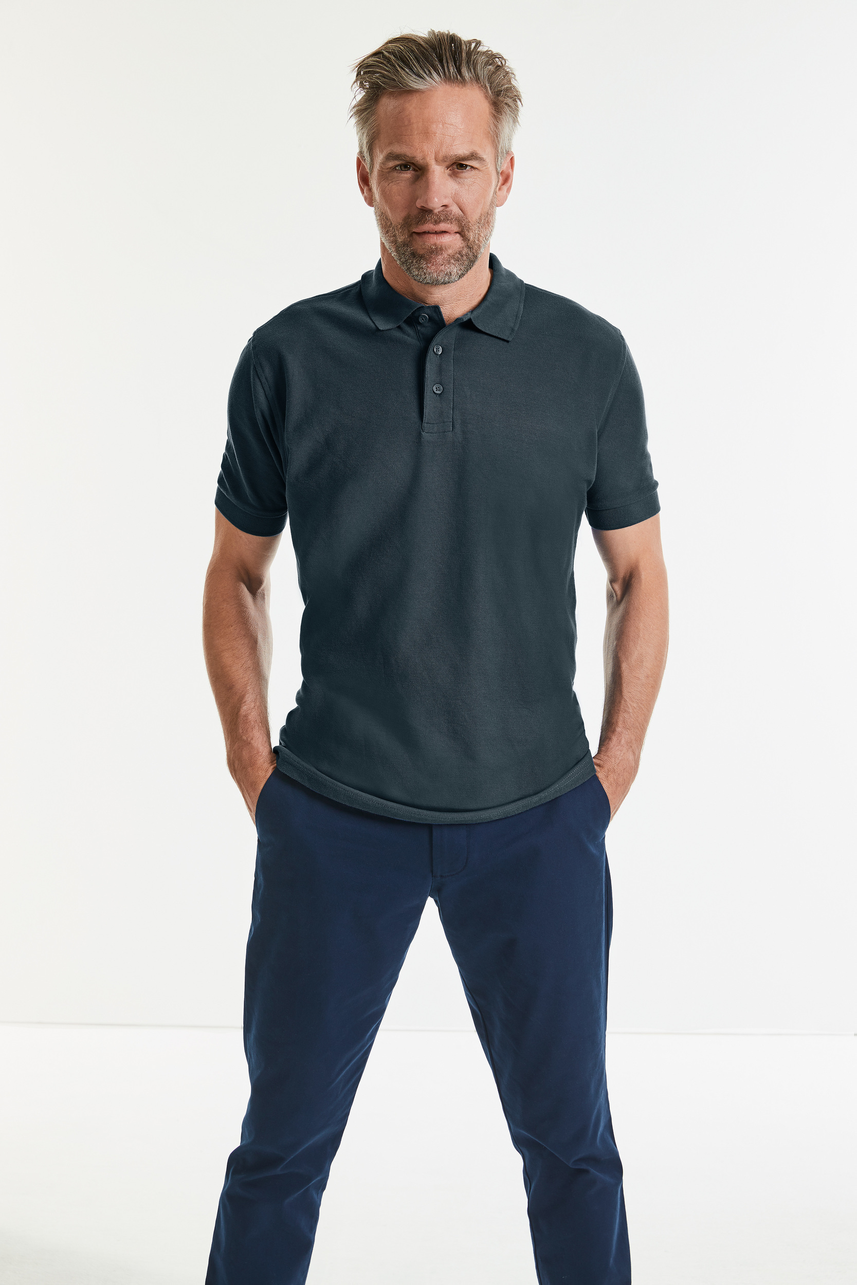 Hombre de pie con polo azul marino y pantalón oscuro, manos en los bolsillos sobre fondo blanco