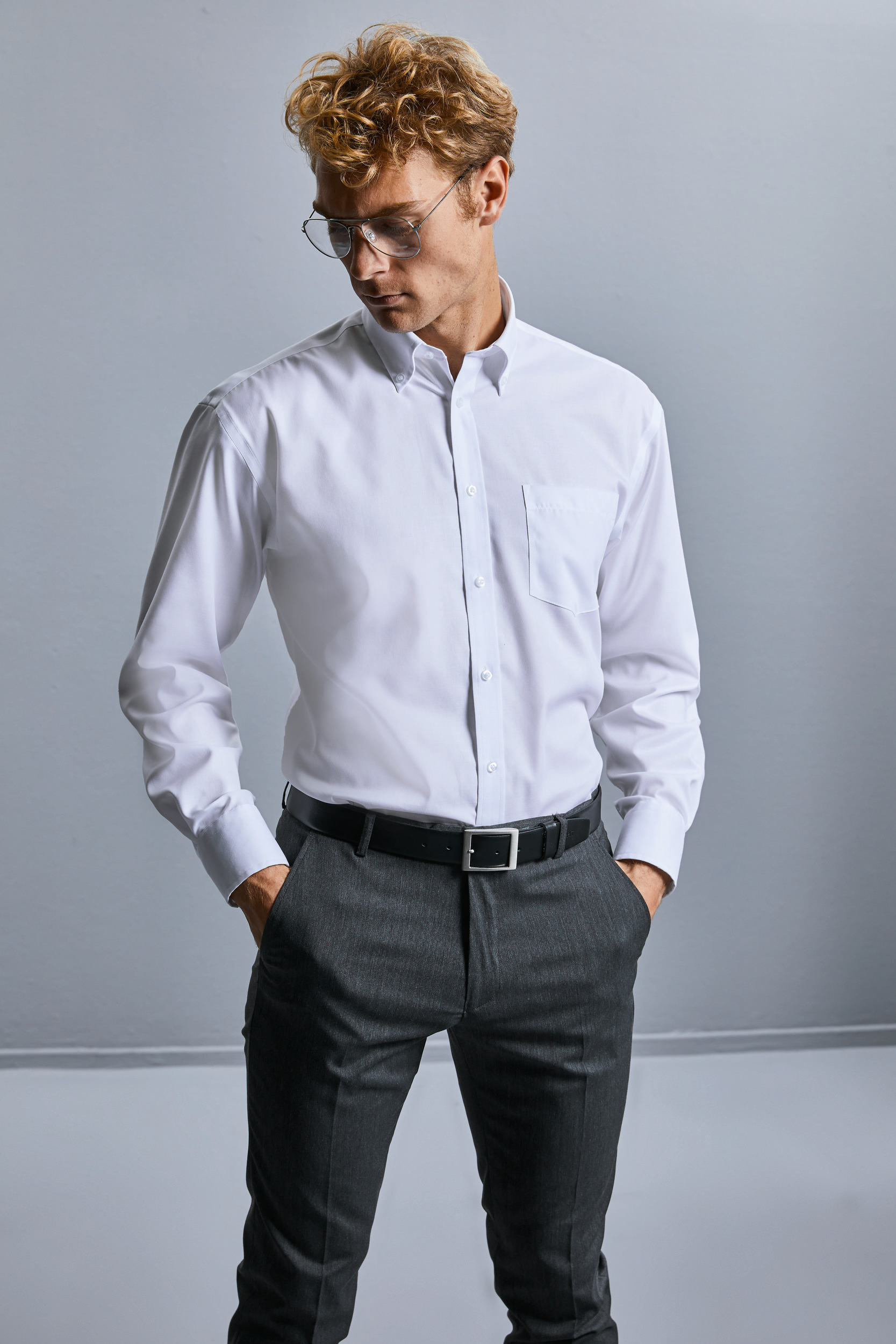 Hombre con camisa blanca de vestir y pantalones grises de traje con las manos en los bolsillos mirando hacia abajo