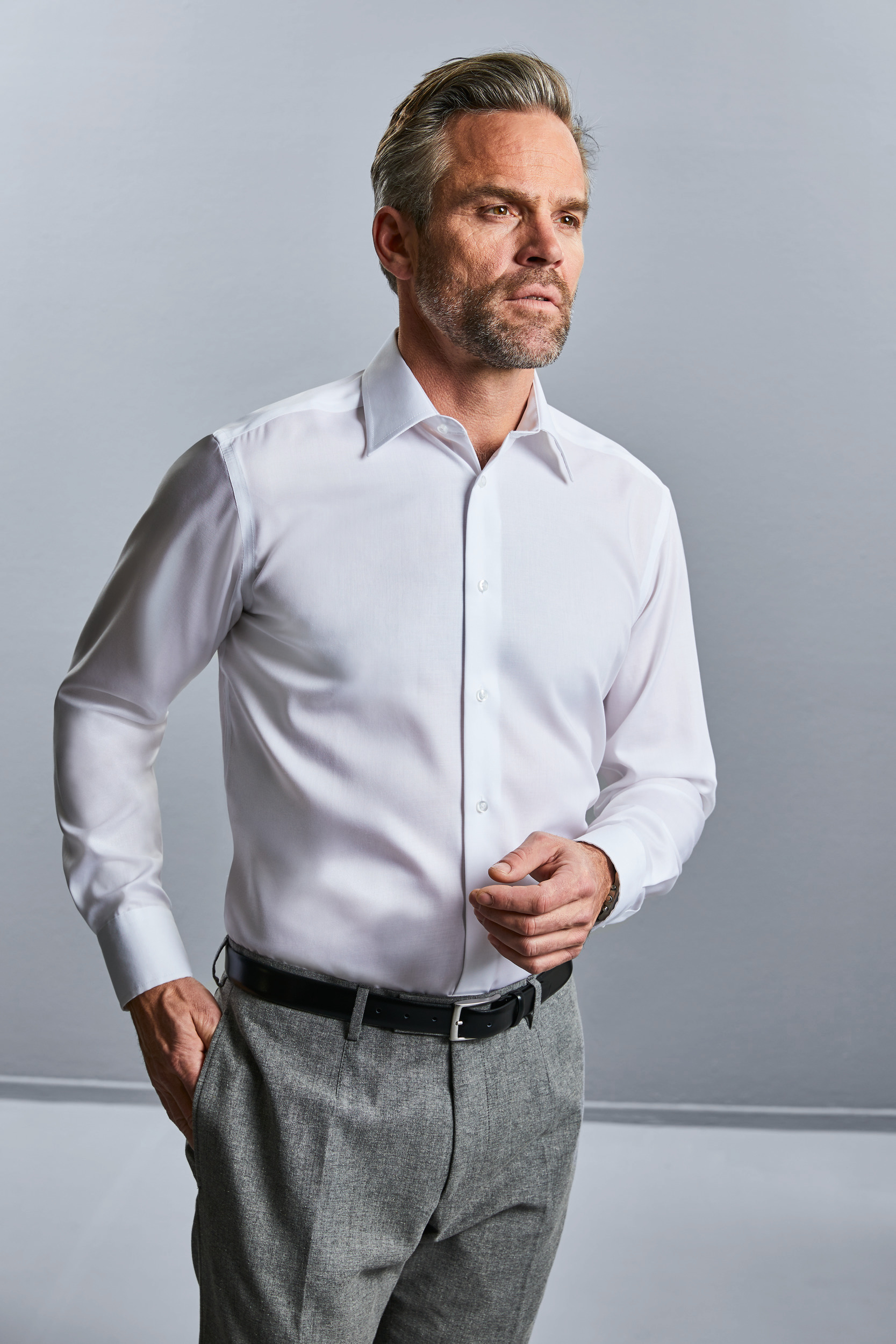 Hombre con camisa blanca y pantalón gris en fondo neutro, posando de perfil.