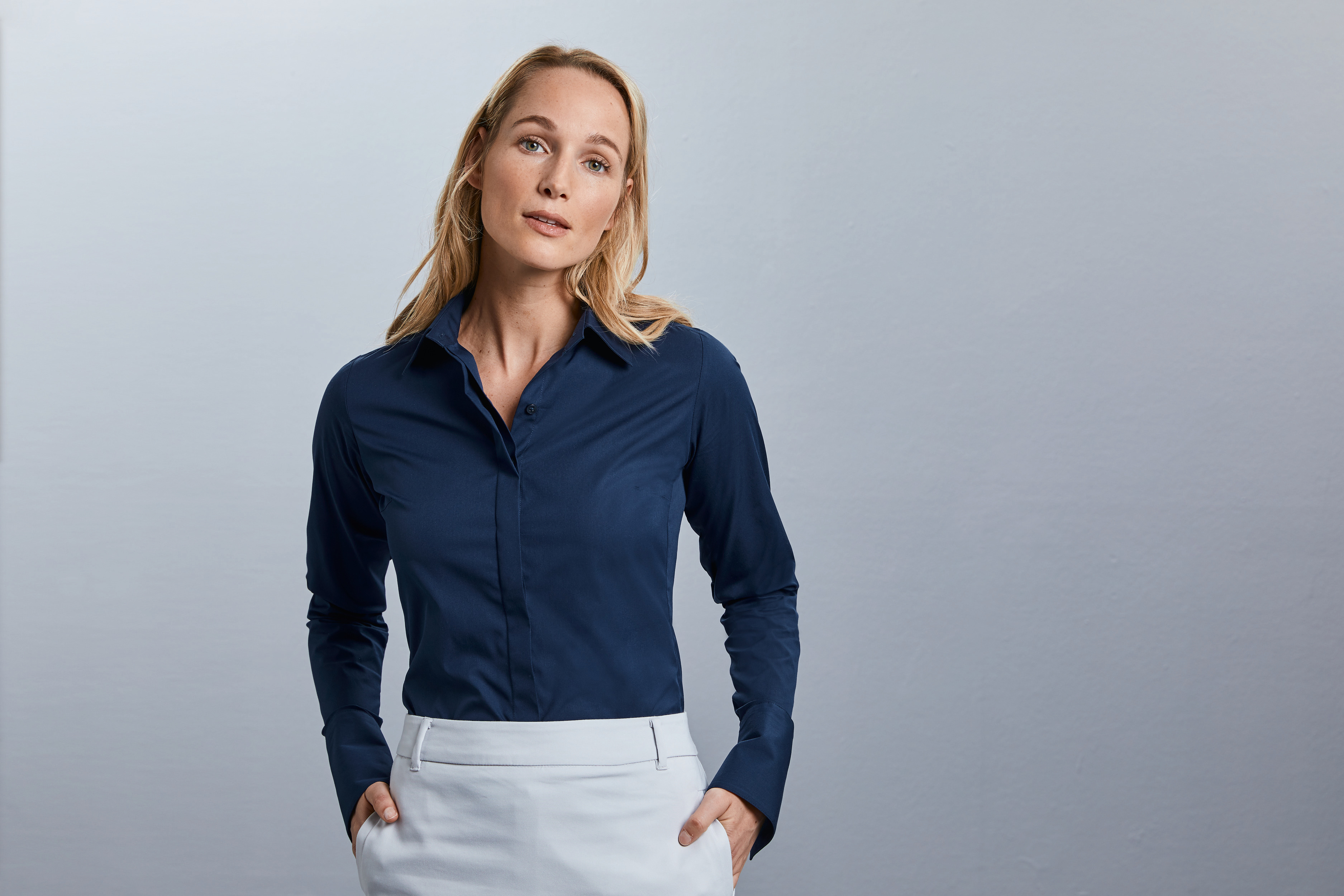 Mujer rubia con camisa azul marino de manga larga y falda blanca sobre fondo gris claro