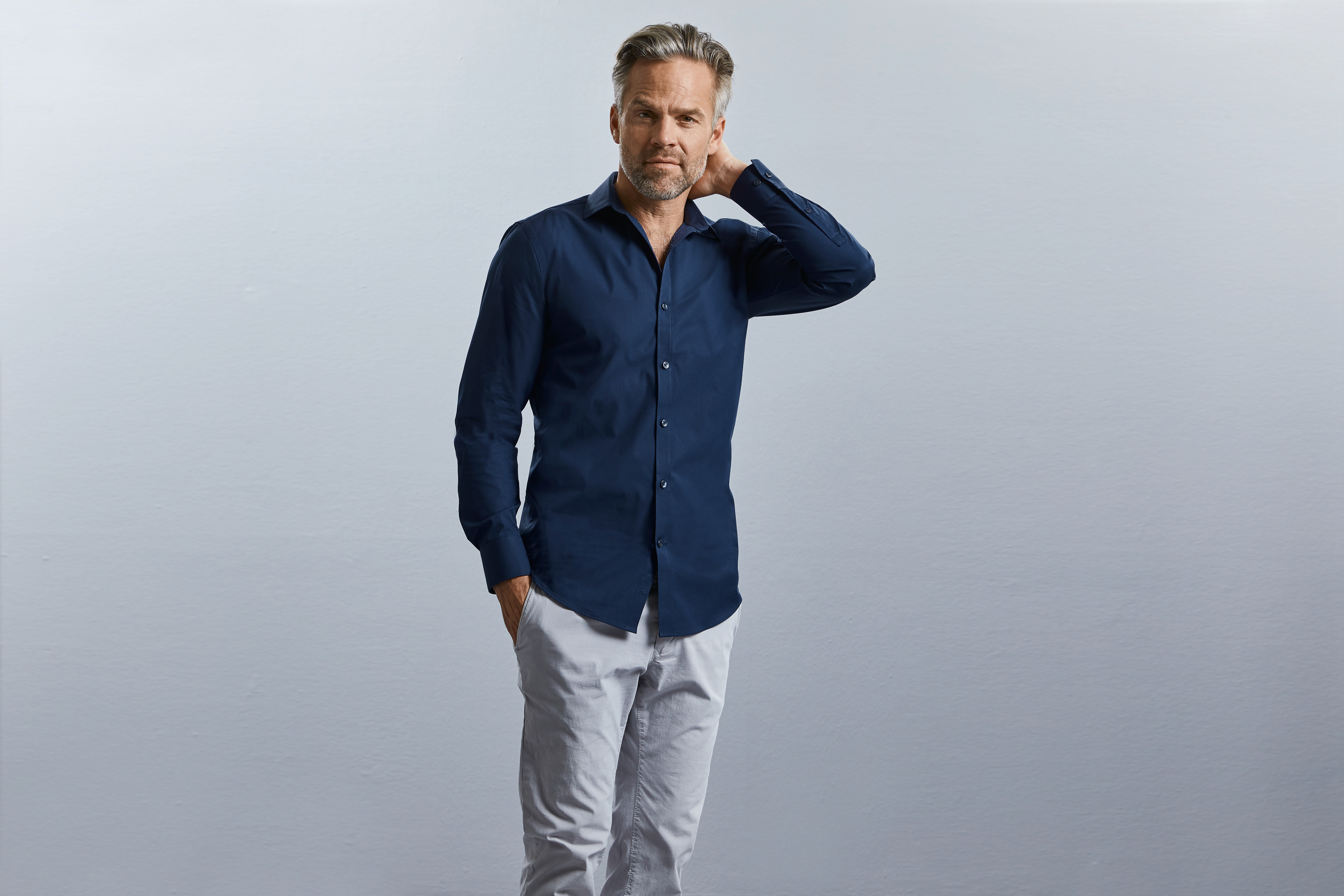 Hombre con camisa azul marino y pantalones claros en un fondo gris, posando con la mano en el cuello.