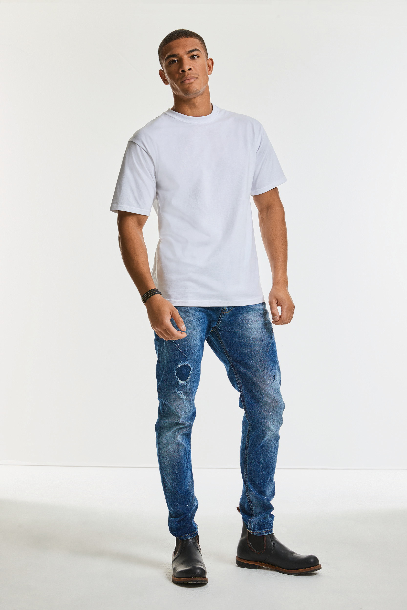 Hombre joven de pie con camiseta blanca básica, vaqueros desgastados y botas negras.