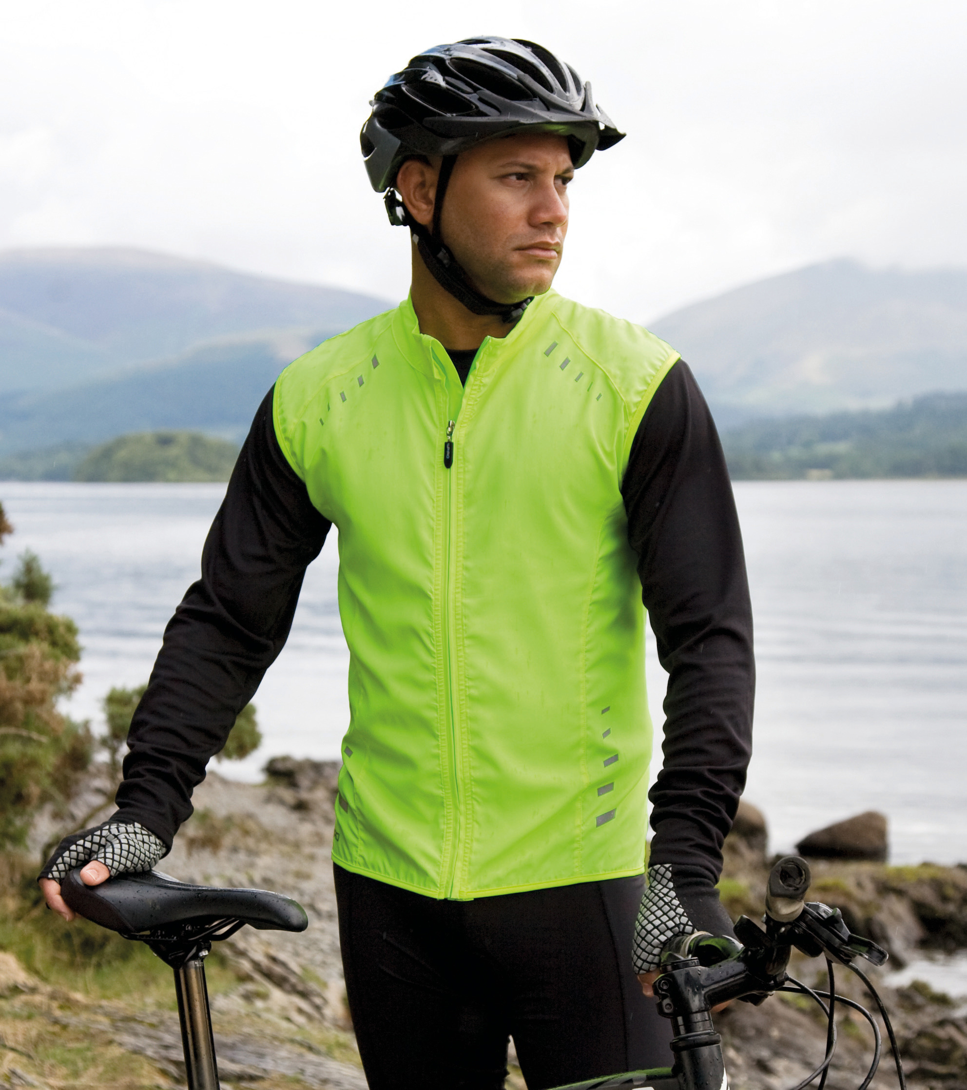 Hombre con casco y chaleco reflectante verde junto a su bicicleta en la orilla de un lago en un paisaje montañoso.