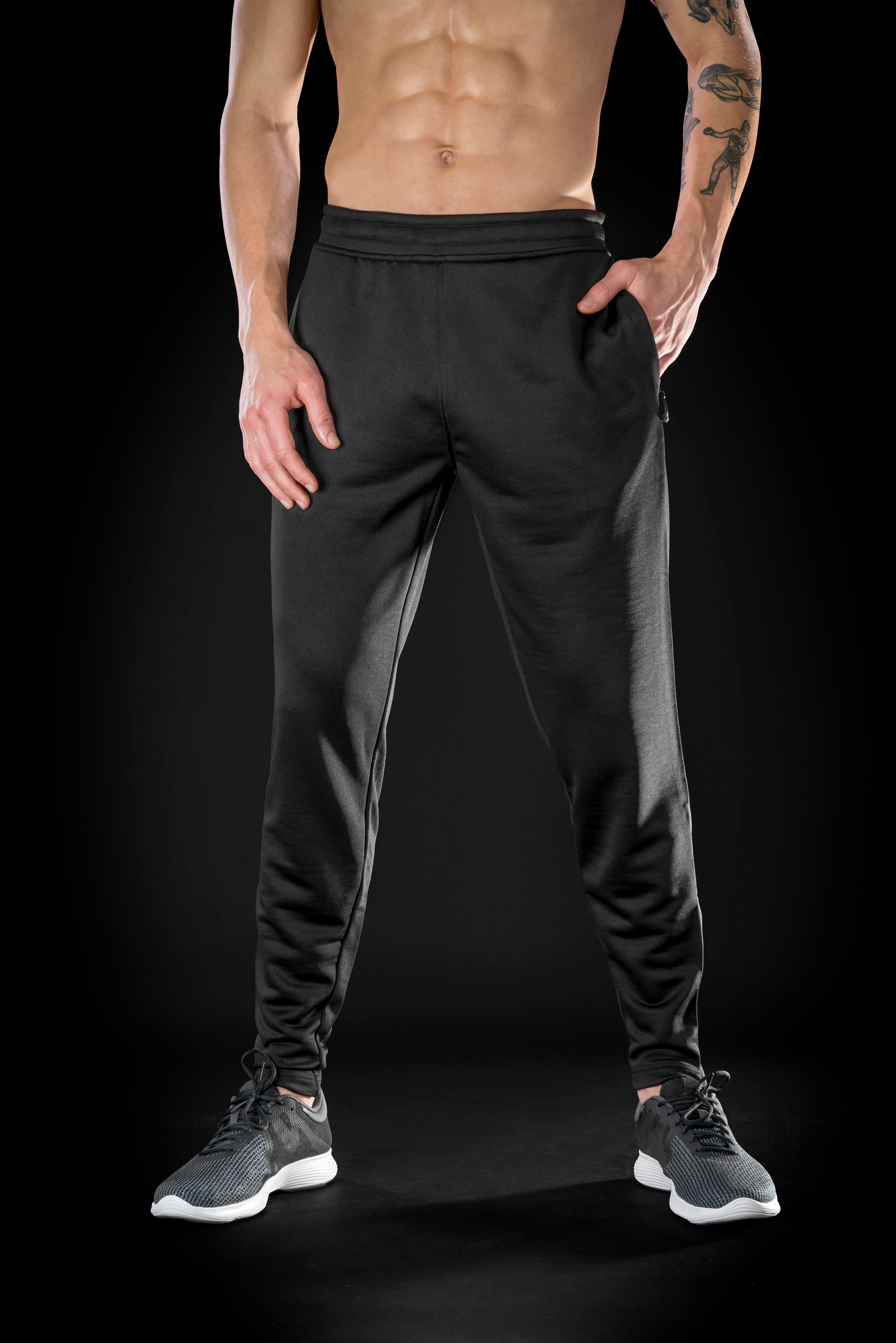 Modelo masculino sin camiseta con pantalones deportivos negros y zapatillas grises sobre fondo oscuro.