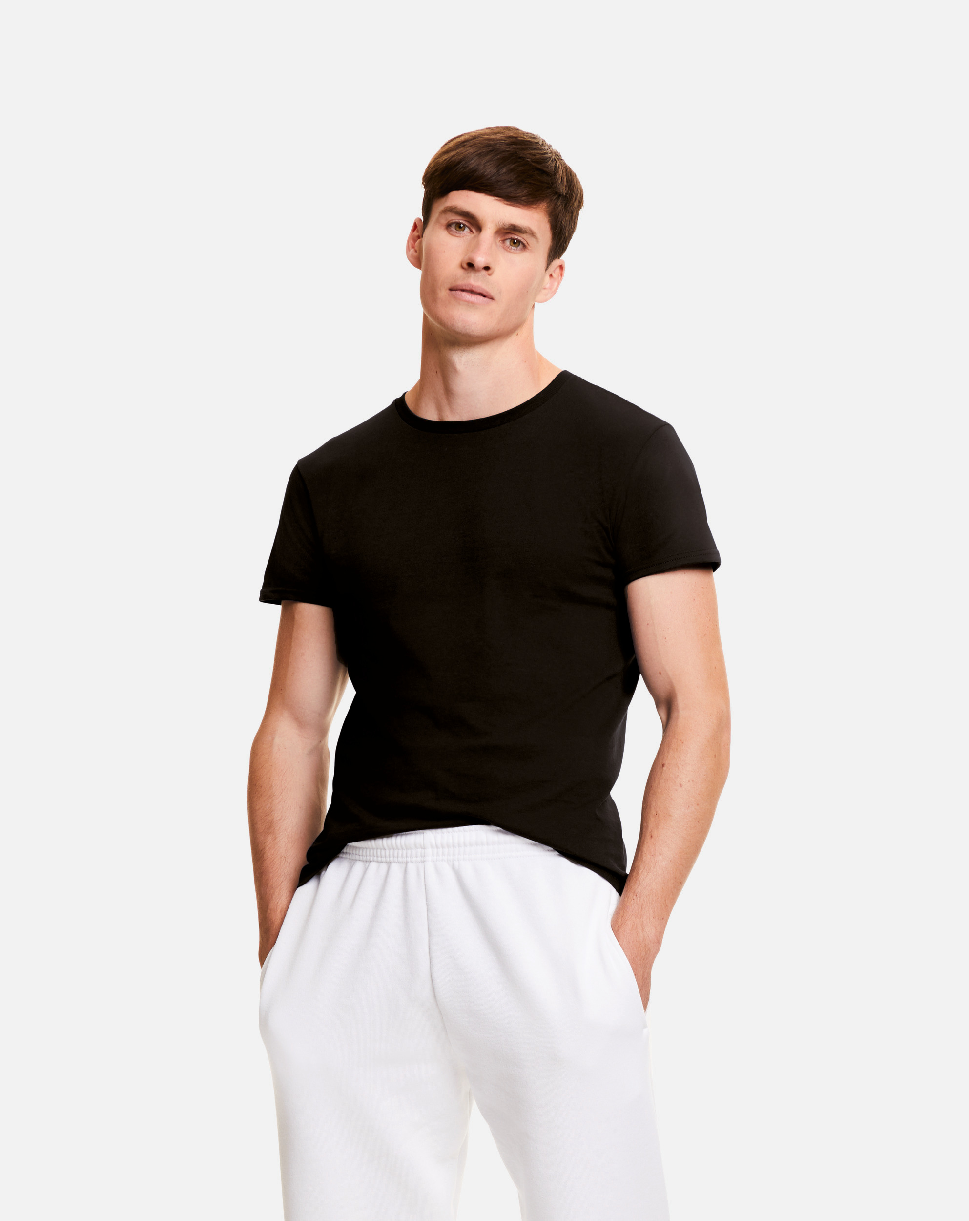 Hombre joven de pie con camiseta negra de manga corta y pantalones deportivos blancos sobre fondo claro.