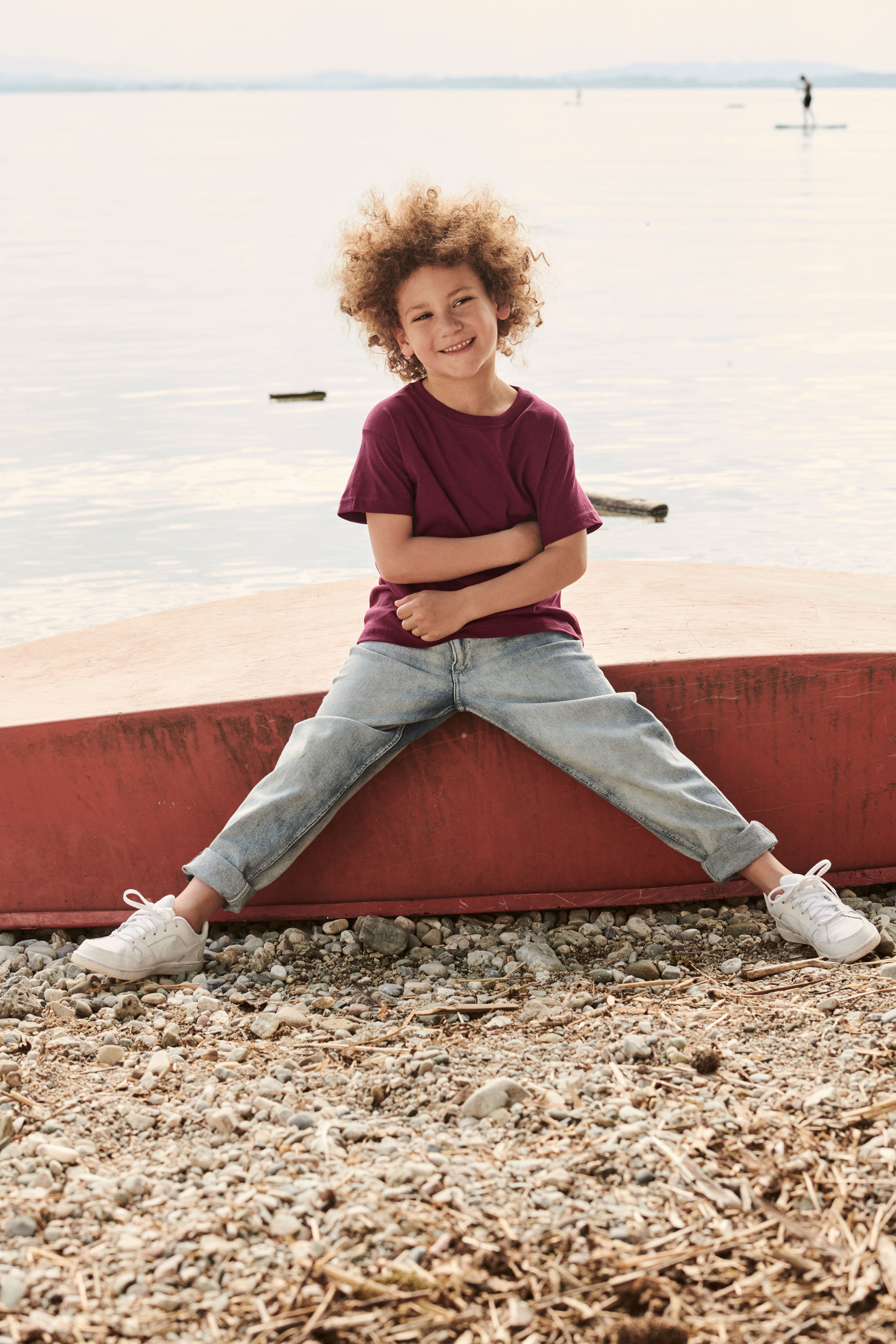 niño sonriente con camiseta burdeos y vaqueros sentado sobre un barco rojo volcado en la orilla de un lago de agua tranquila