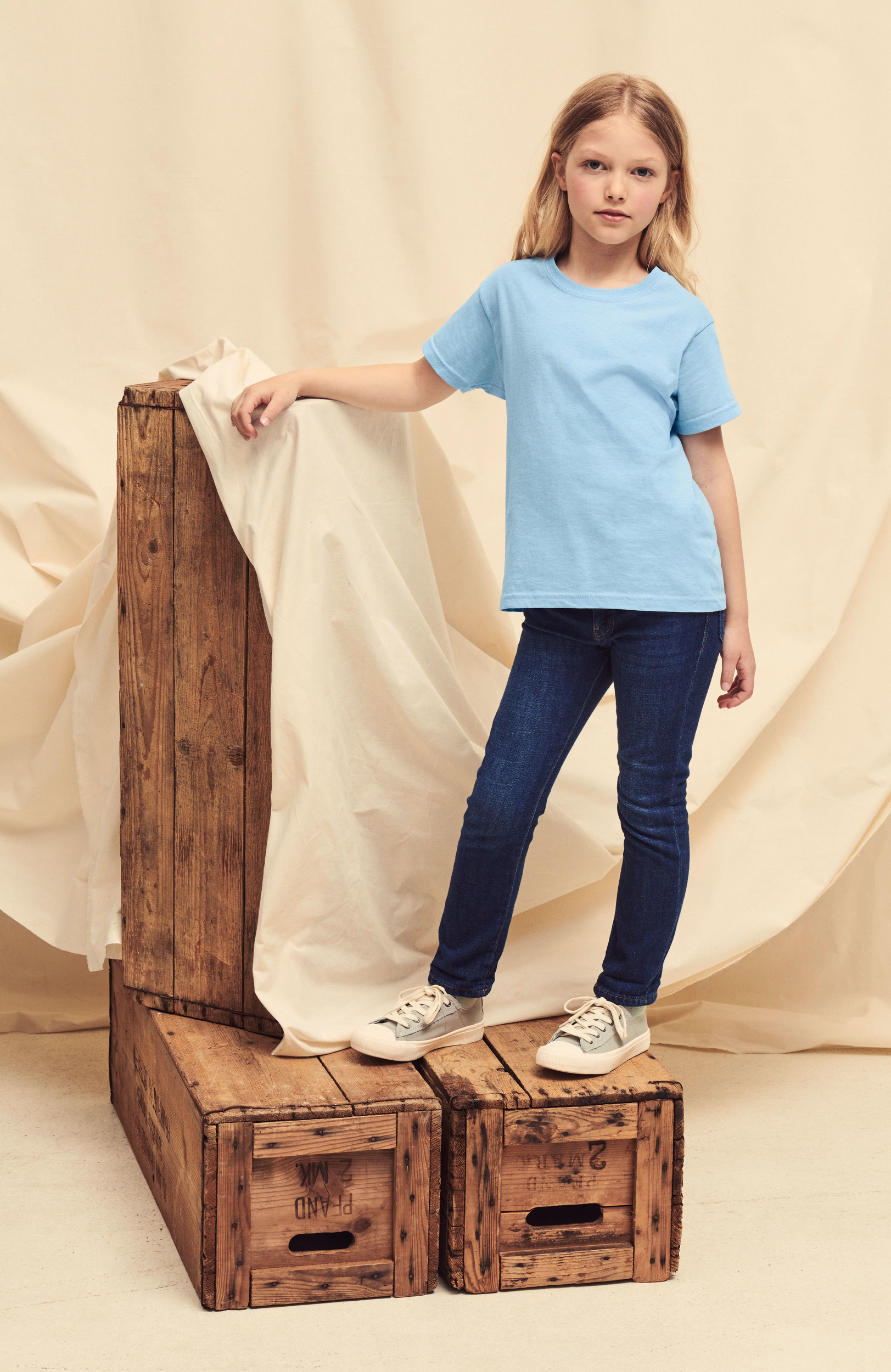 Niña rubia de pie sobre cajas de madera con camiseta azul y vaqueros ante un fondo de tela drapeada
