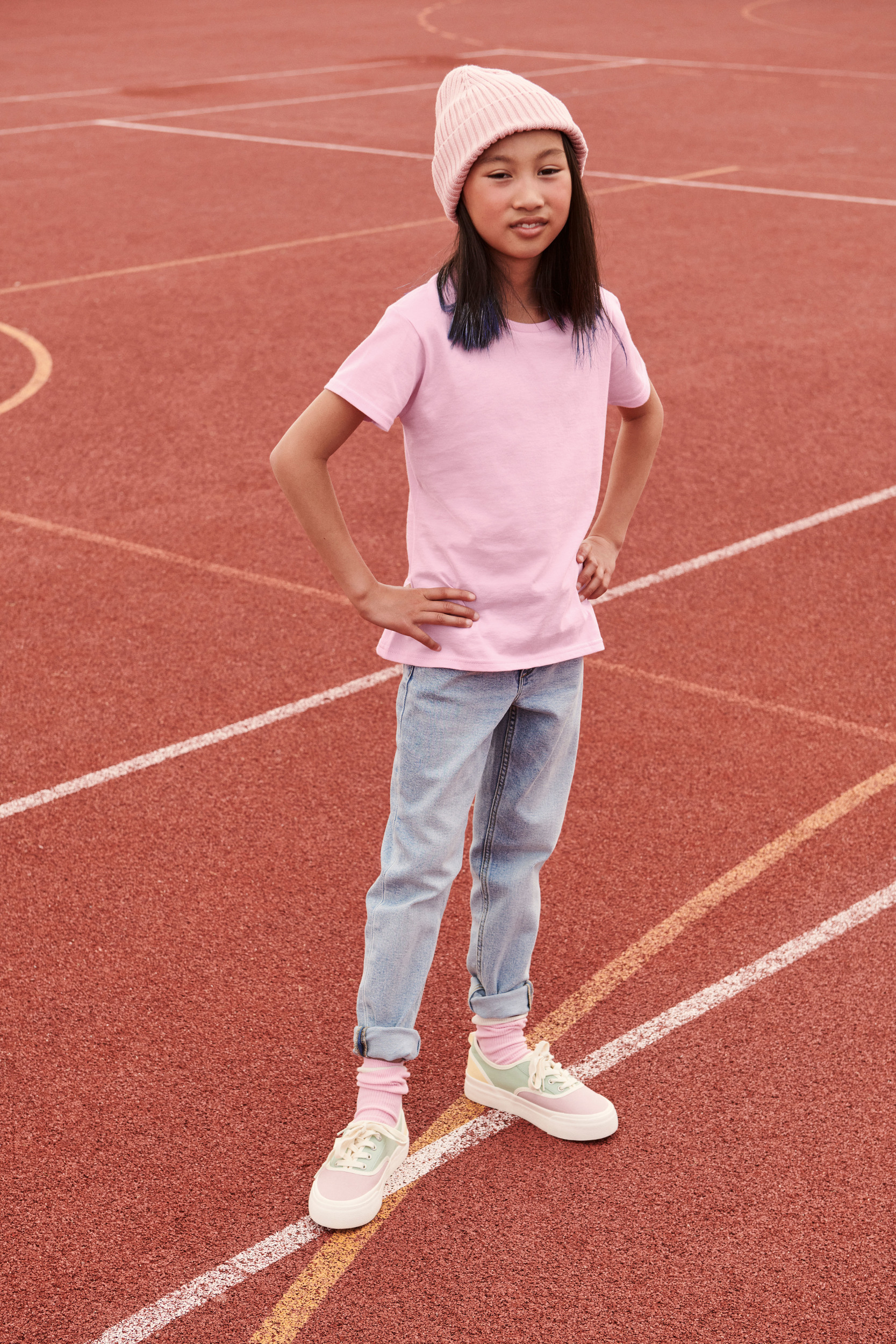 Niña con camiseta y gorro rosa de pie con las manos en la cintura sobre una pista de atletismo roja