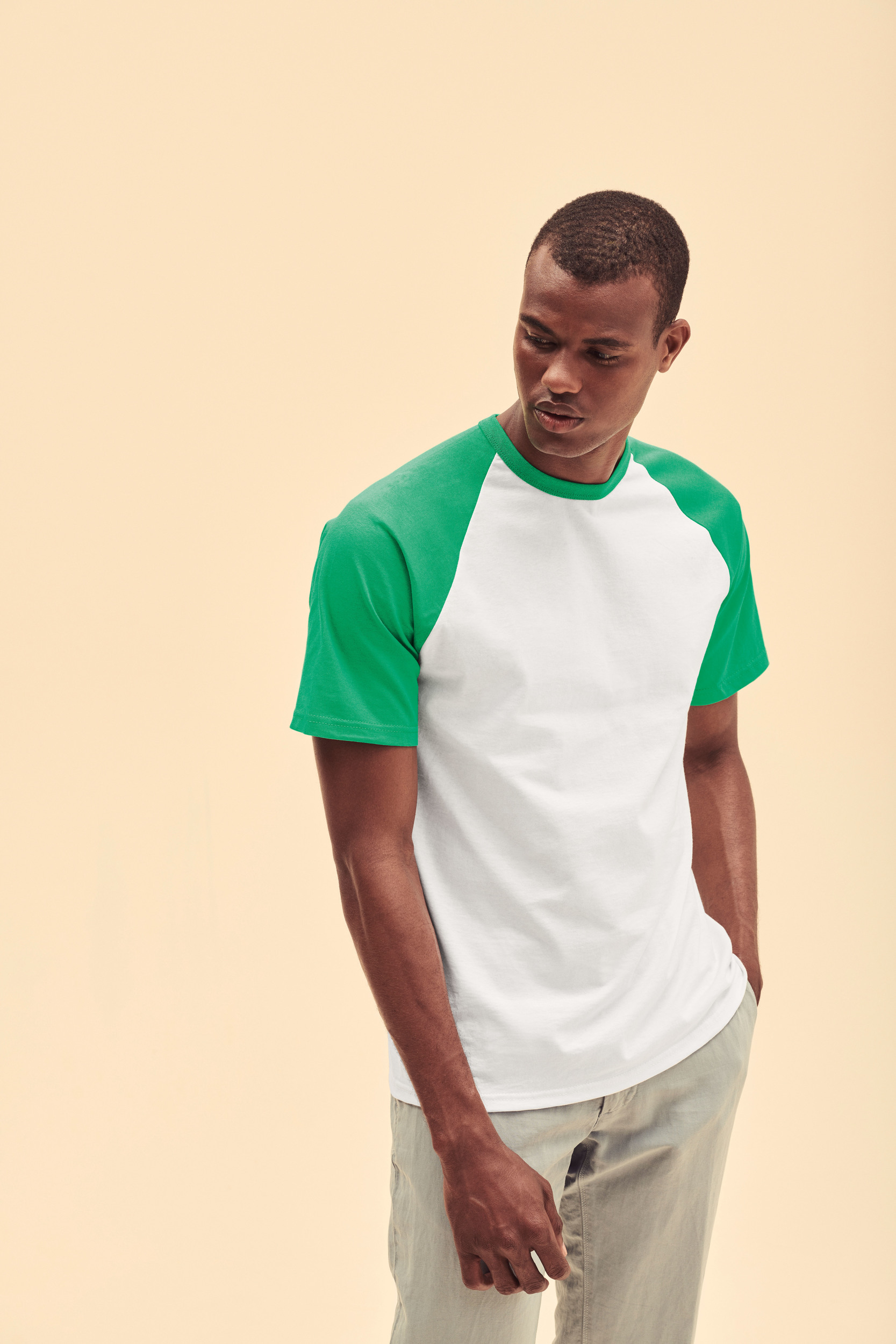 hombre joven de pie con camiseta blanca de mangas verdes y pantalón claro sobre fondo beige claro
