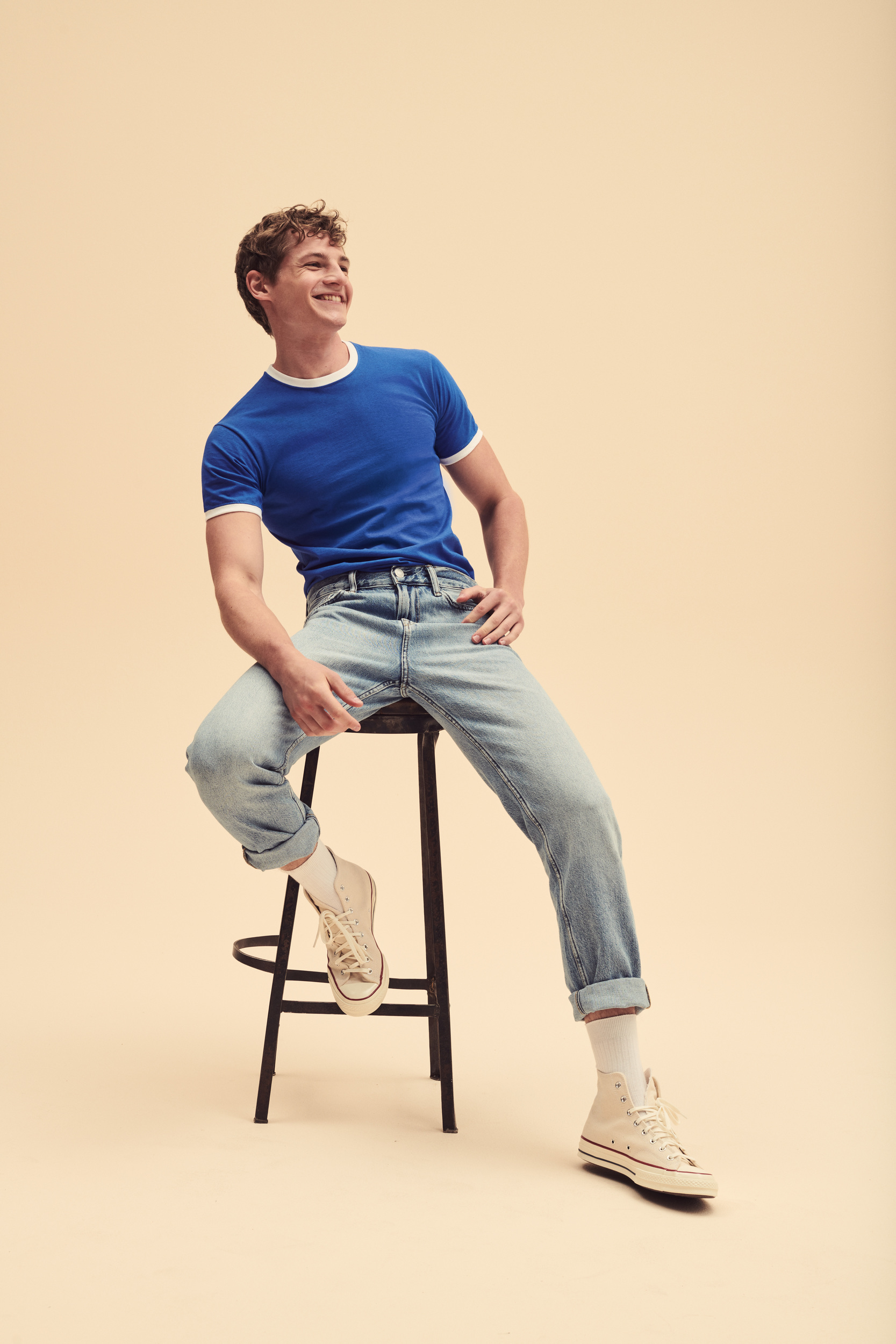 Hombre joven sonriente sentado en un taburete con camiseta azul, vaqueros y zapatillas sobre fondo beige