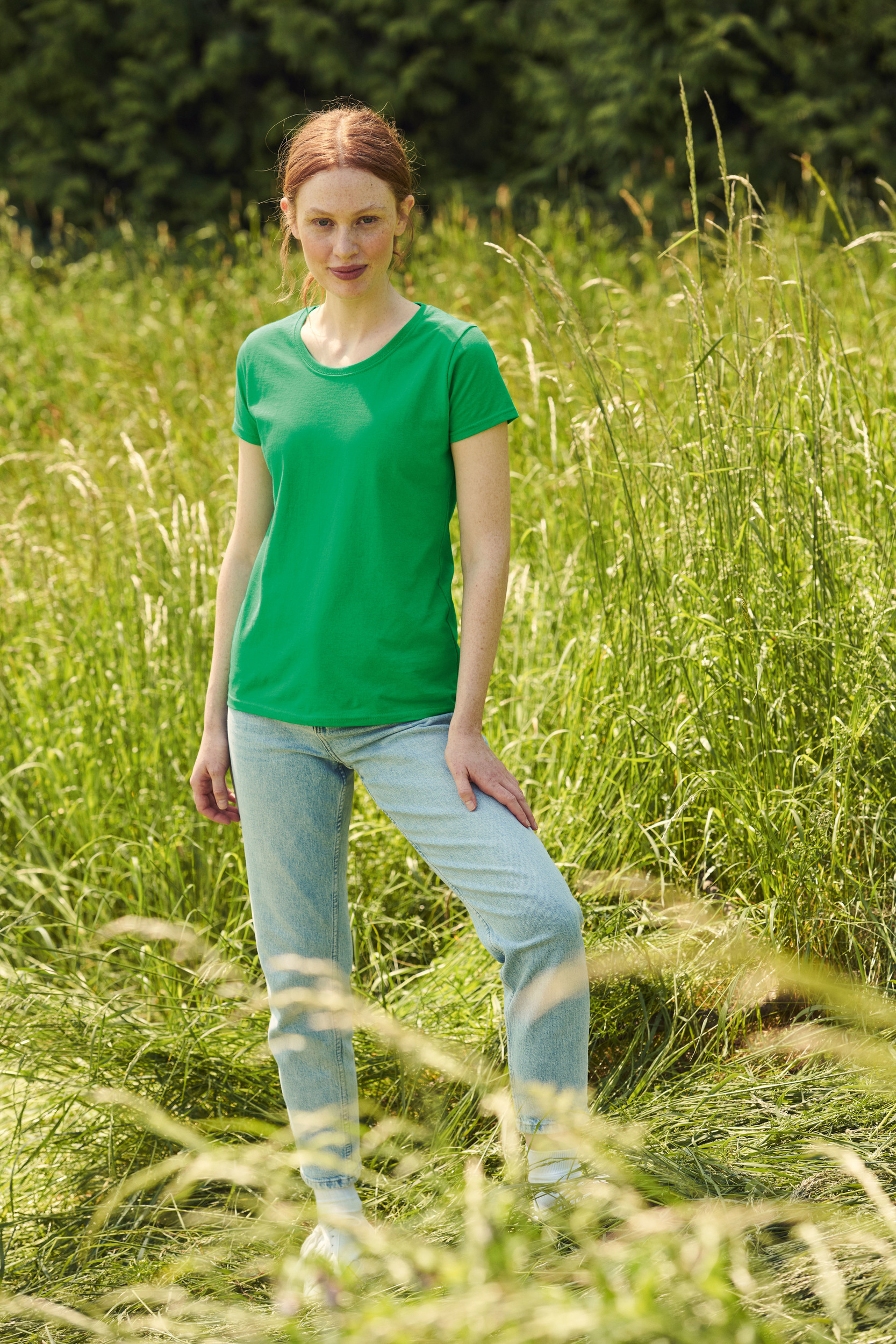 Mujer pelirroja con camiseta verde y vaqueros de pie en un campo de hierba alta.