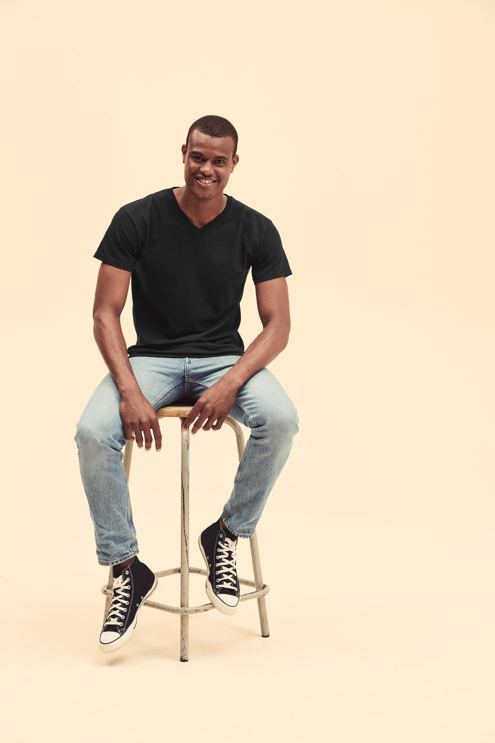 Hombre joven sonriente sentado en un taburete sobre fondo liso, con camiseta negra, vaqueros azul claro y zapatillas negras de caña alta.