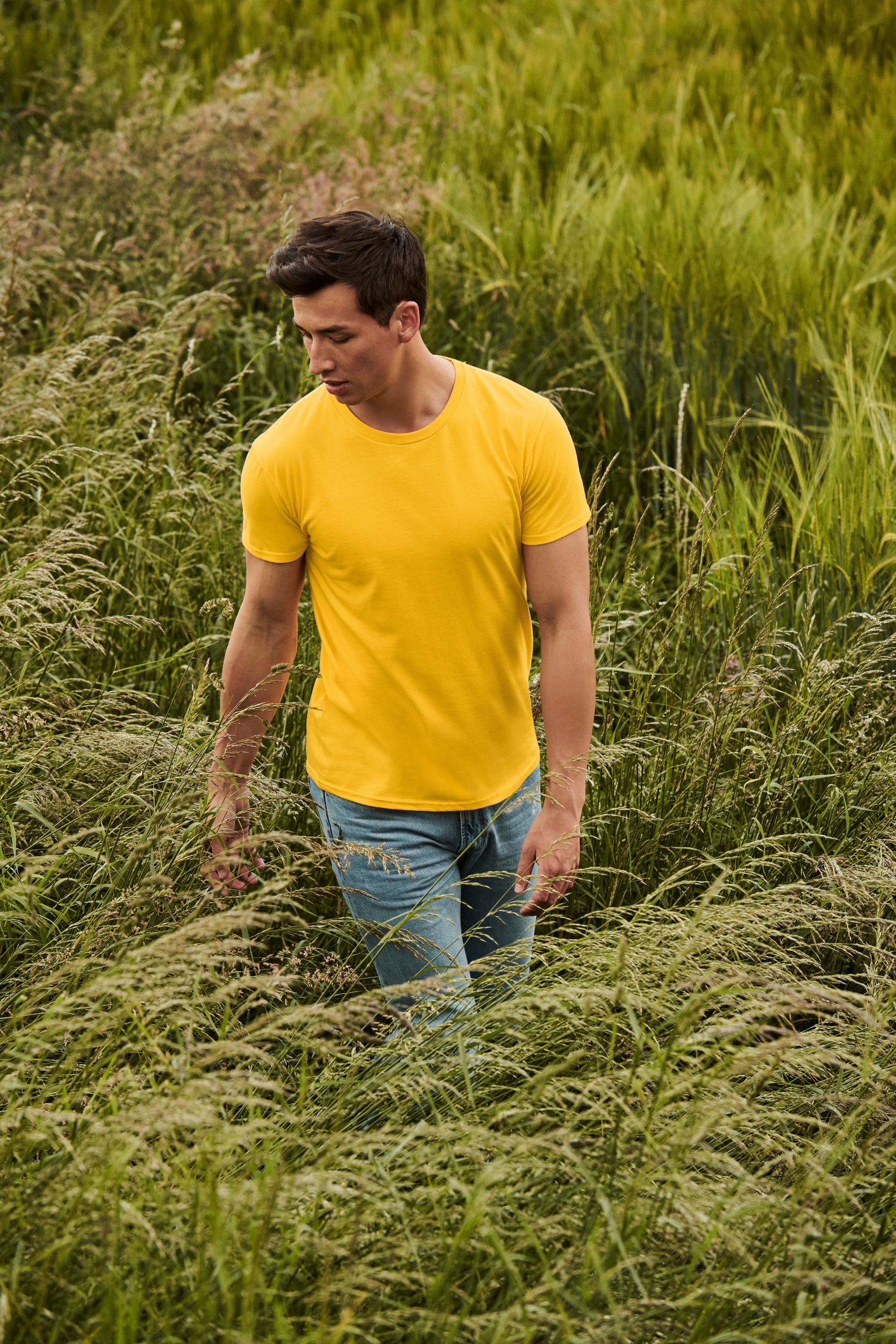 Hombre con camiseta amarilla y vaqueros paseando entre altas hierbas en un prado verde.