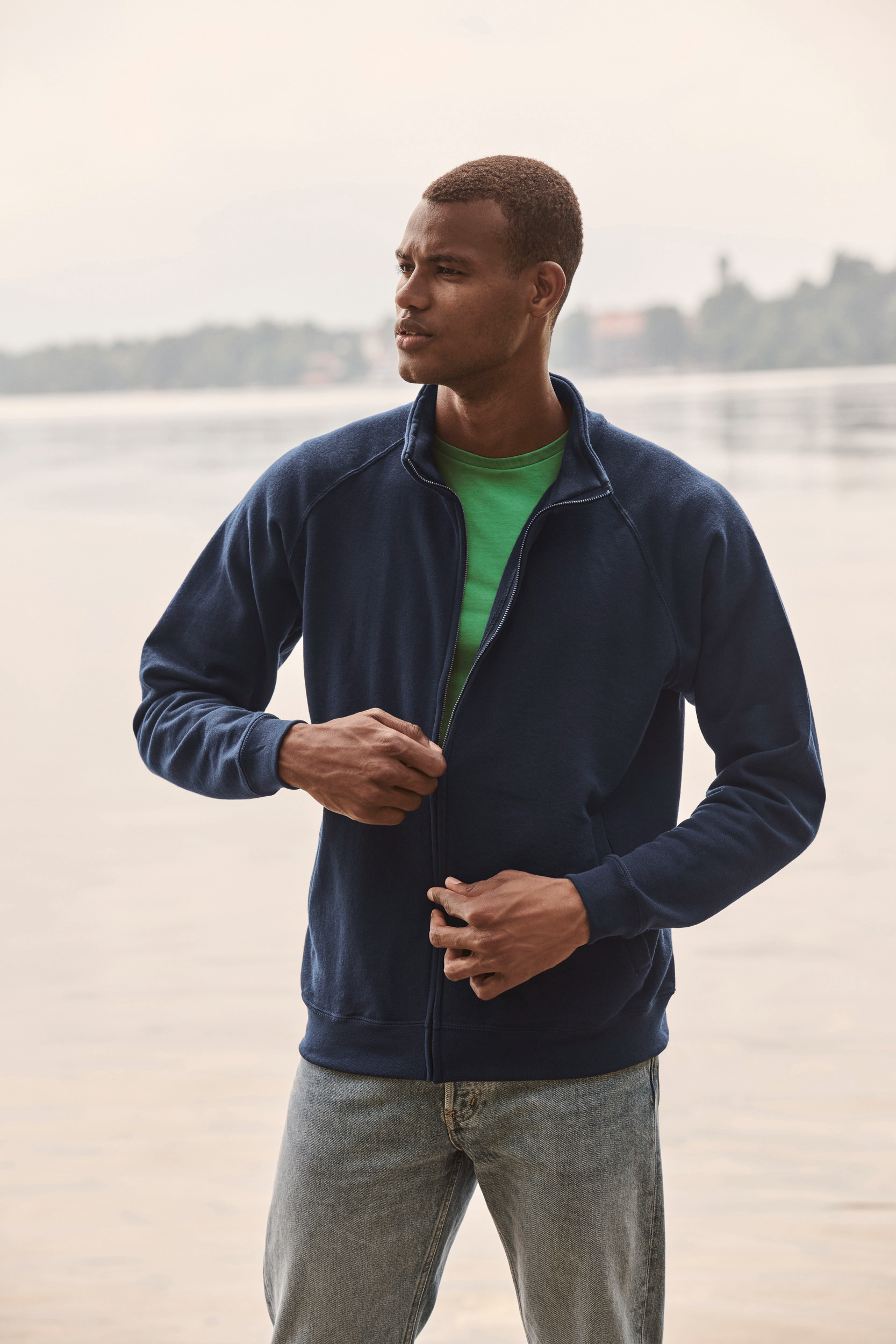 Hombre joven abrochándose una sudadera azul marino junto a un lago al aire libre