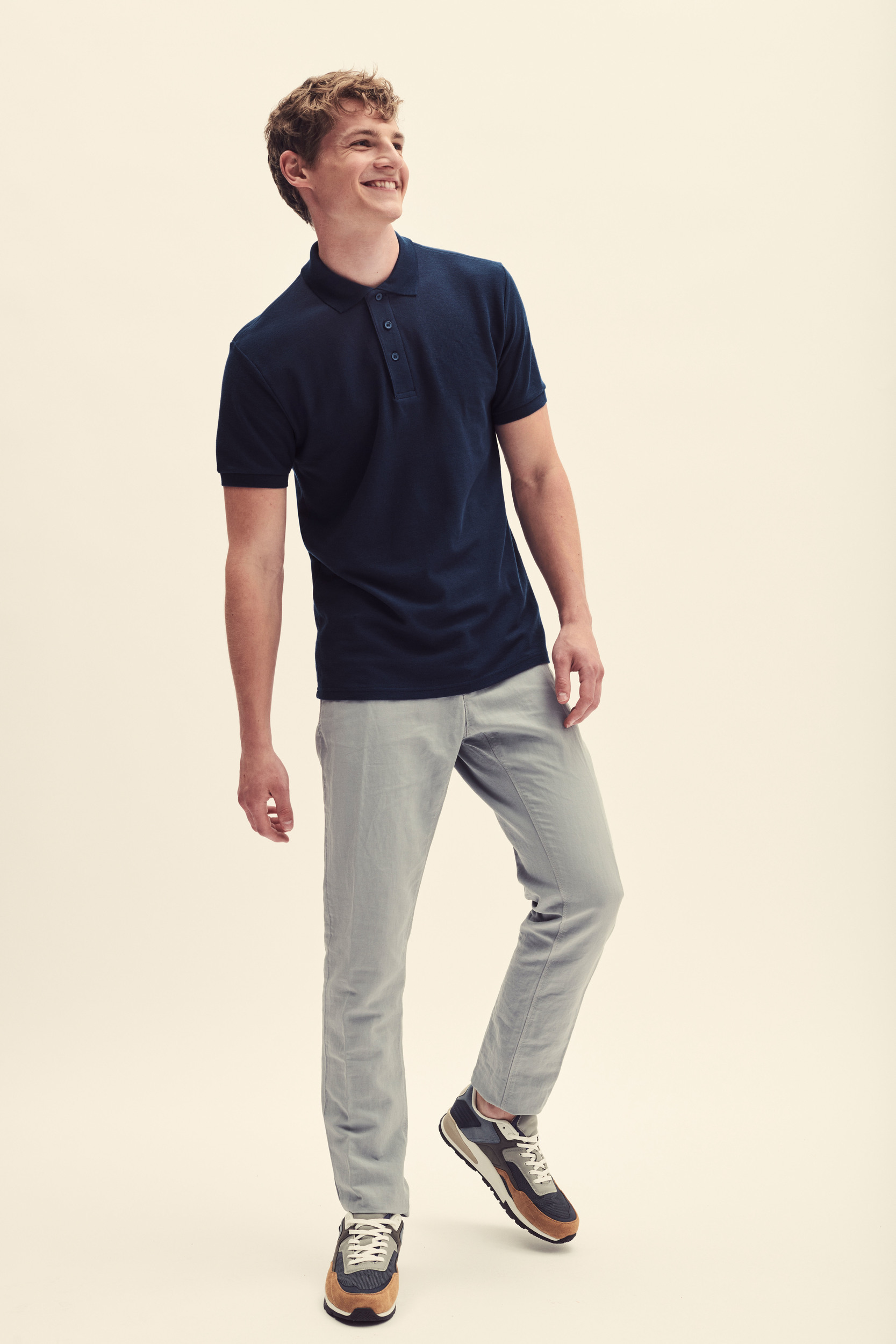 Hombre joven de pie con polo azul marino, pantalones grises y zapatillas casuales sobre fondo neutro