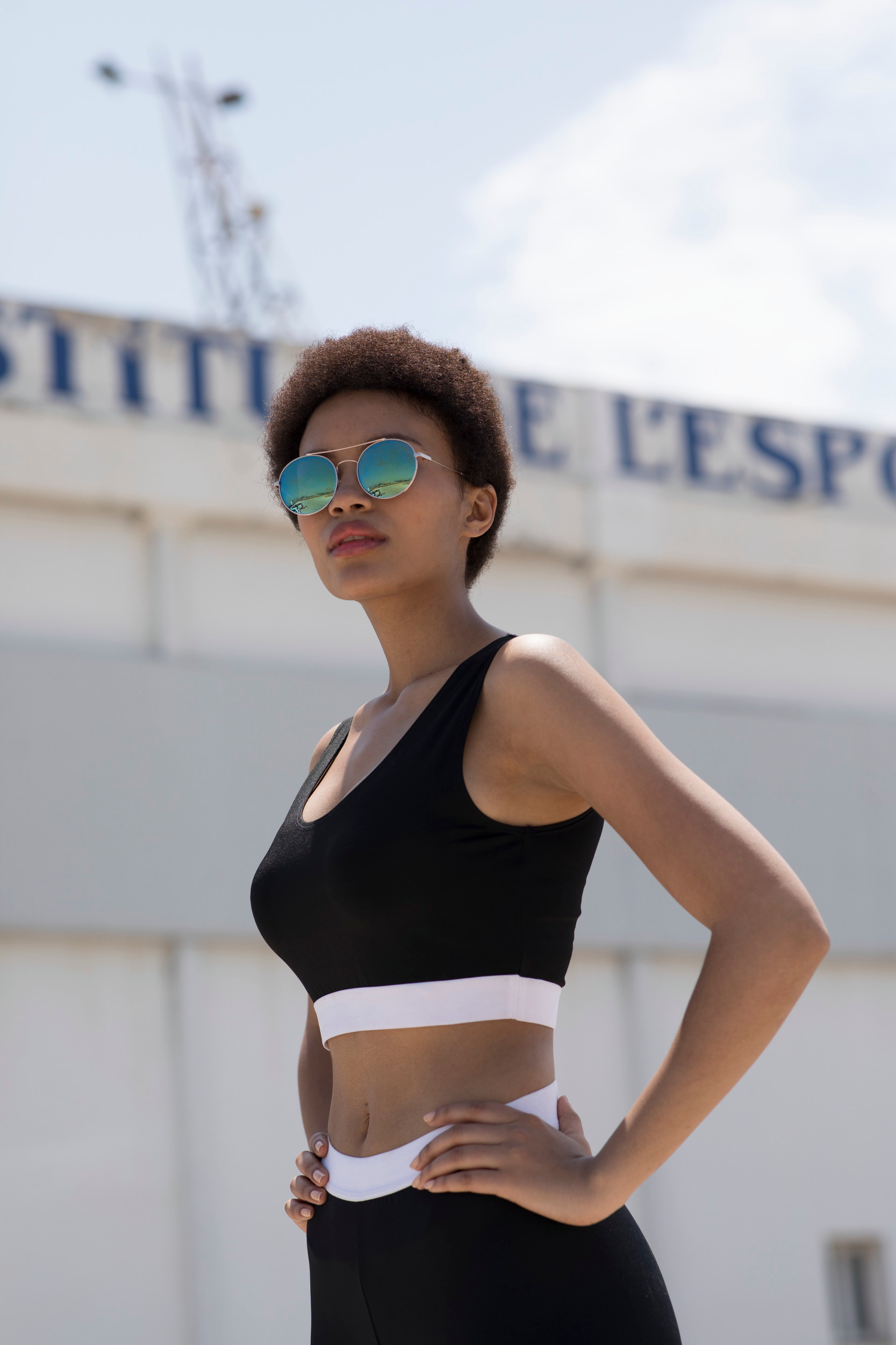 mujer afro con top y mallas deportivas negras posando frente a un edificio deportivo bajo el sol
