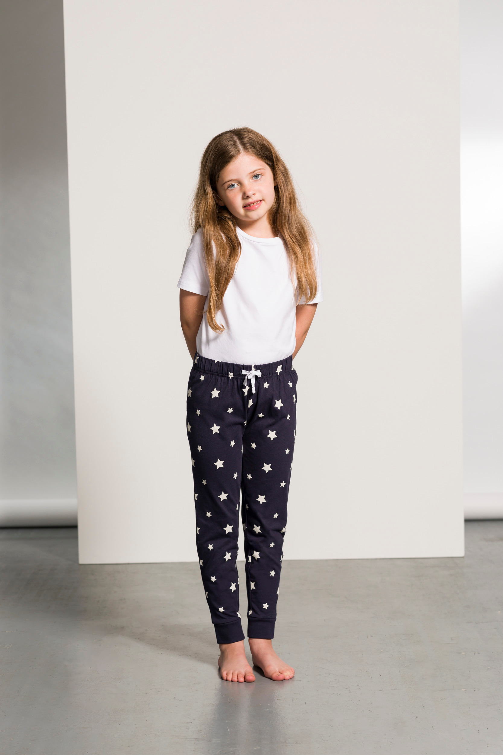 Niña descalza de pie con camiseta blanca y pantalón azul marino estampado de estrellas en un estudio.