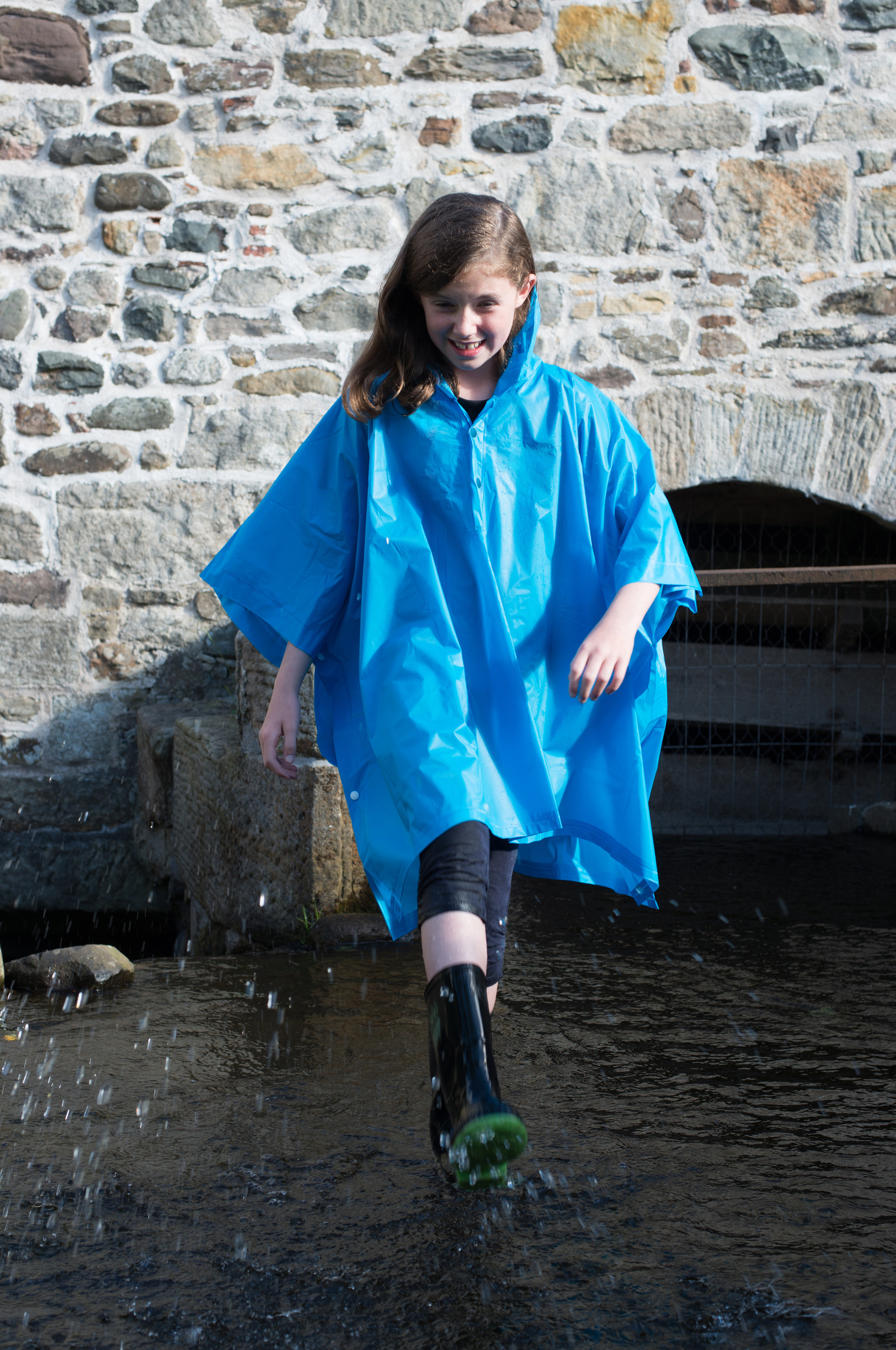 Niña sonriente con chubasquero azul y botas de agua salpicando en un charco junto a un muro de piedra