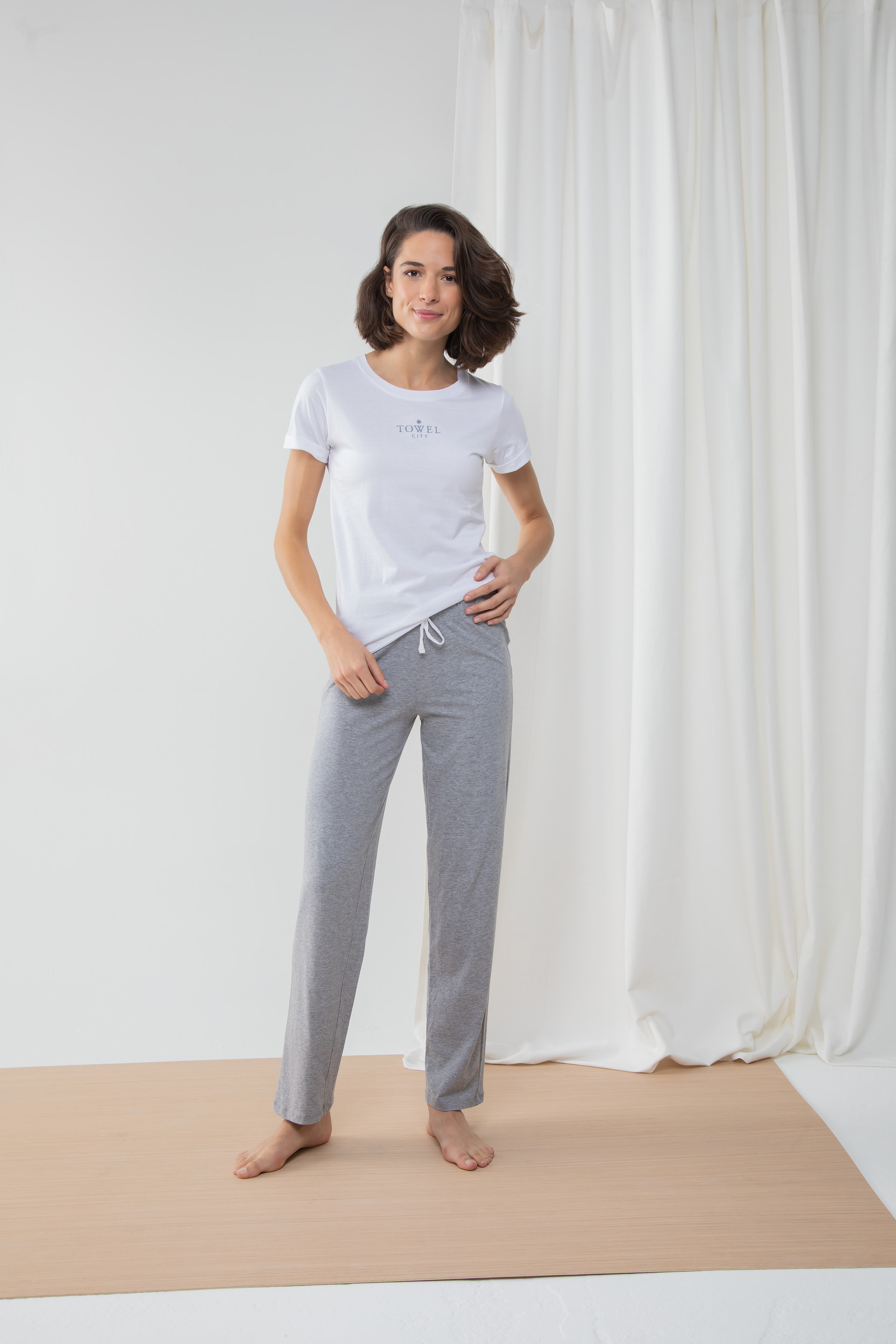 Mujer joven de pie con camiseta blanca y pantalón gris cómodo sobre suelo de madera y fondo de cortina blanca