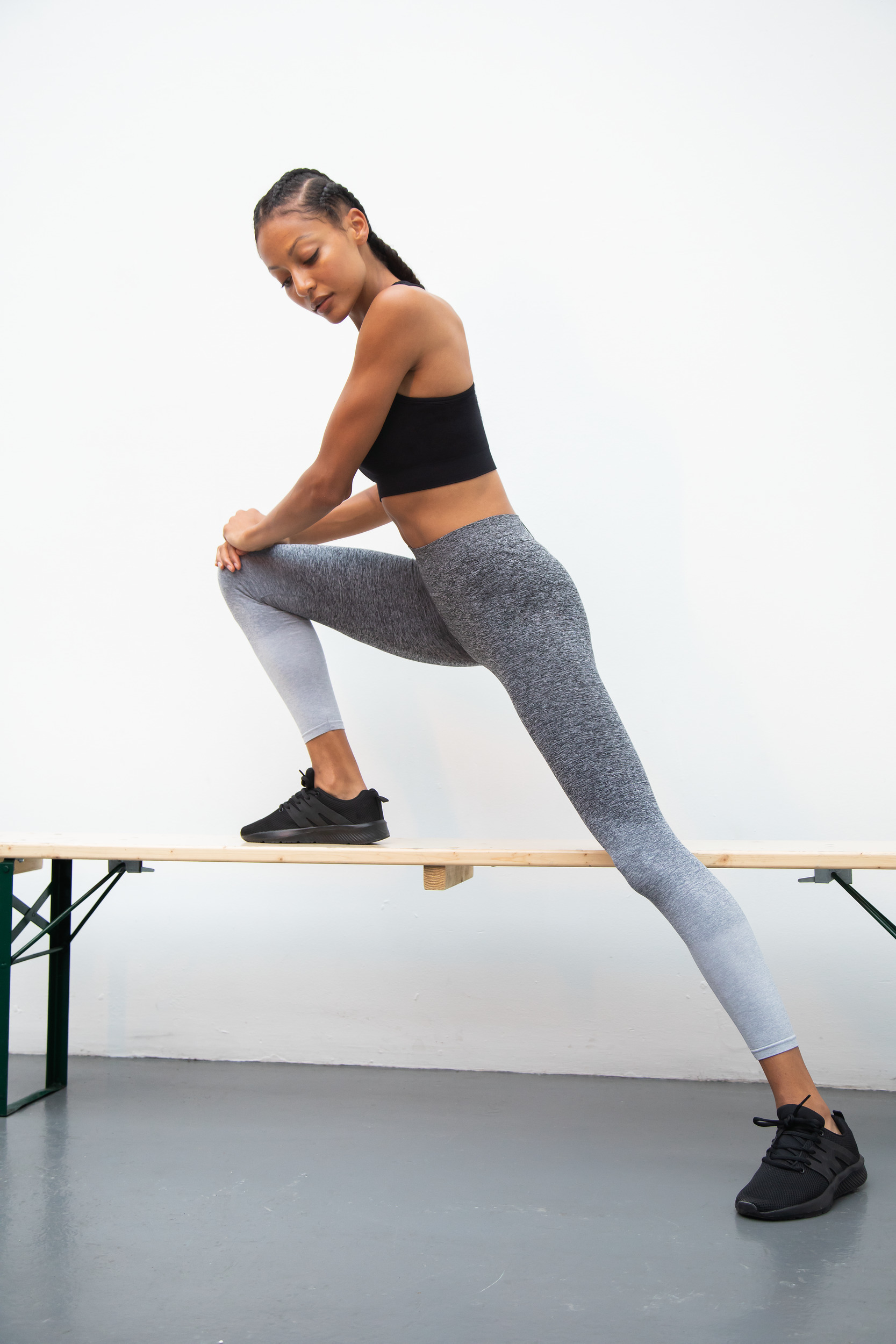 Mujer con ropa deportiva estirando la pierna sobre un banco en un estudio minimalista.