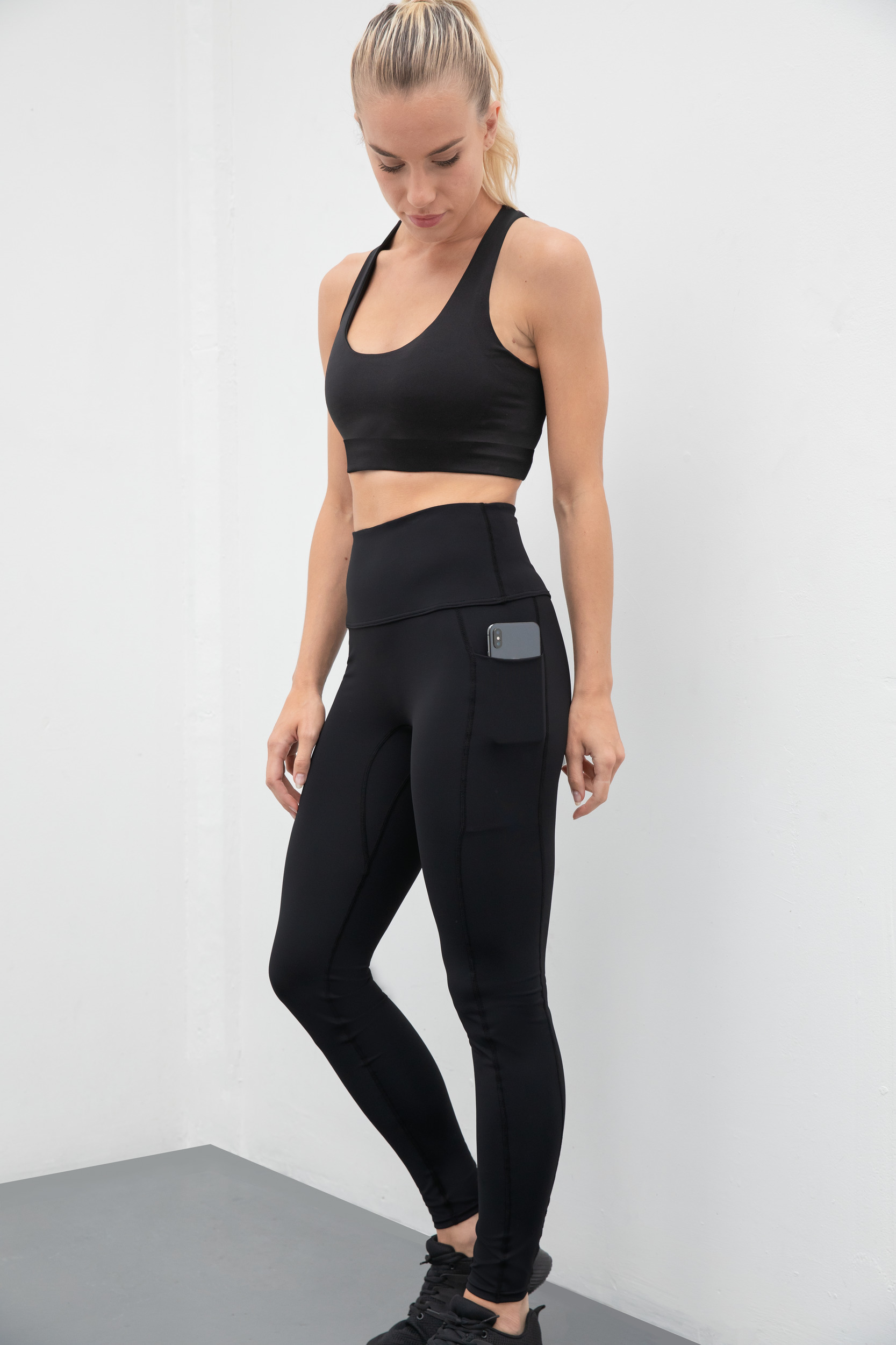 Mujer con top deportivo y mallas negras con un teléfono en el bolsillo lateral, de pie junto a una pared blanca.