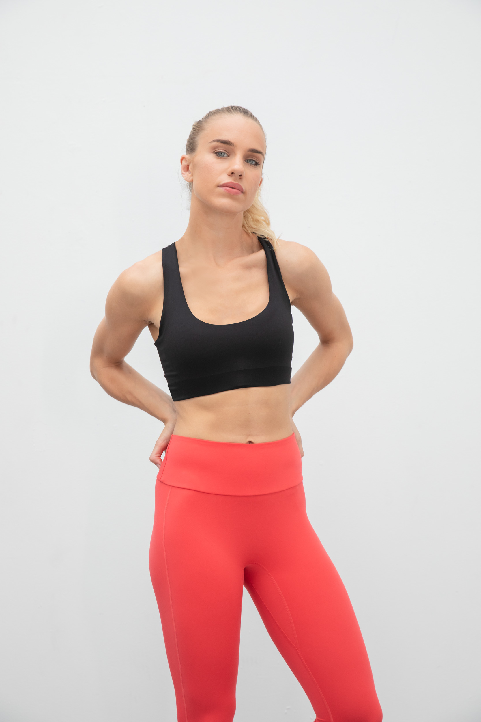 mujer joven con top deportivo negro y mallas rojas de fitness posando de pie sobre fondo blanco