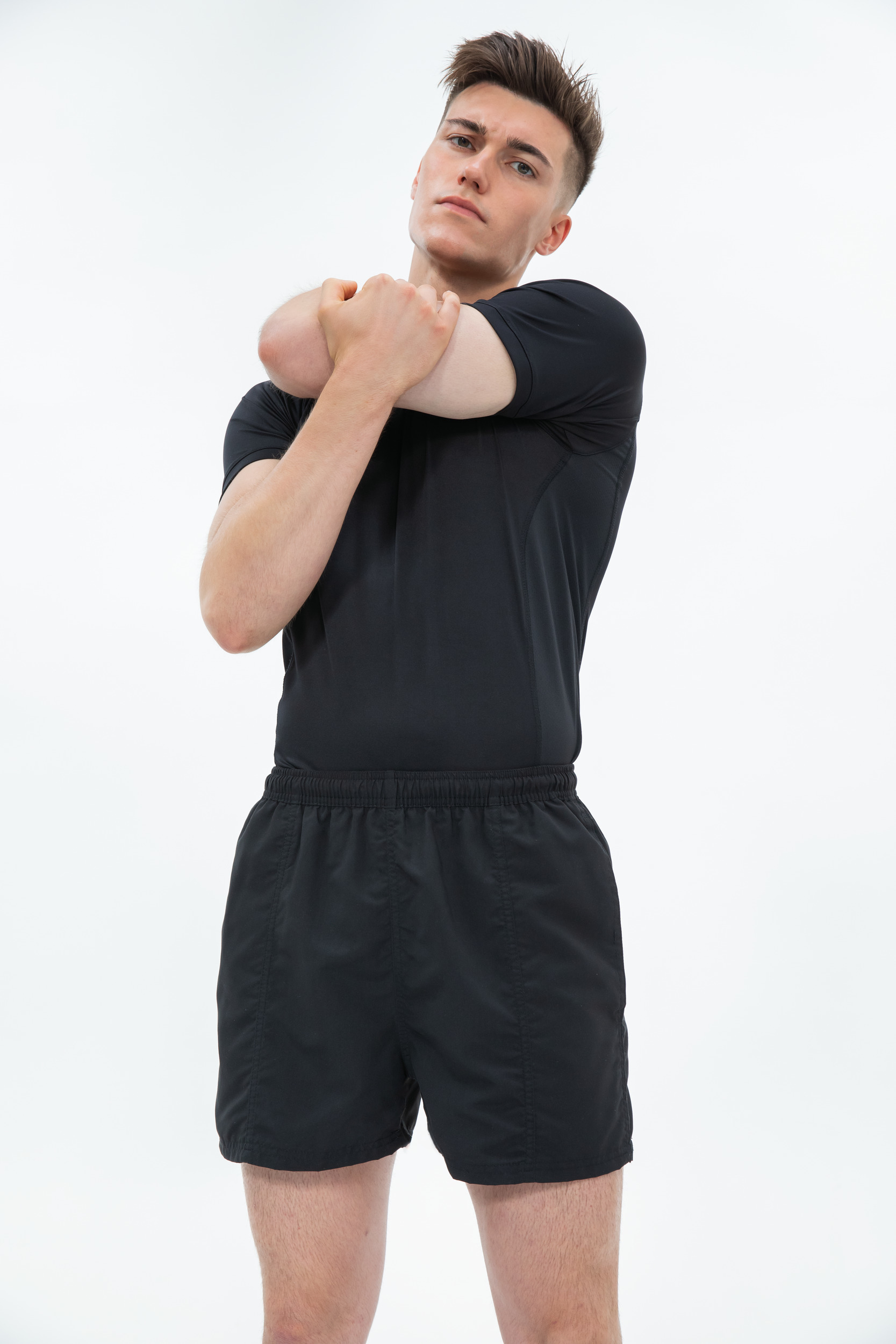 Hombre joven estirando el brazo cruzado al pecho con camiseta y pantalones cortos negros sobre fondo blanco