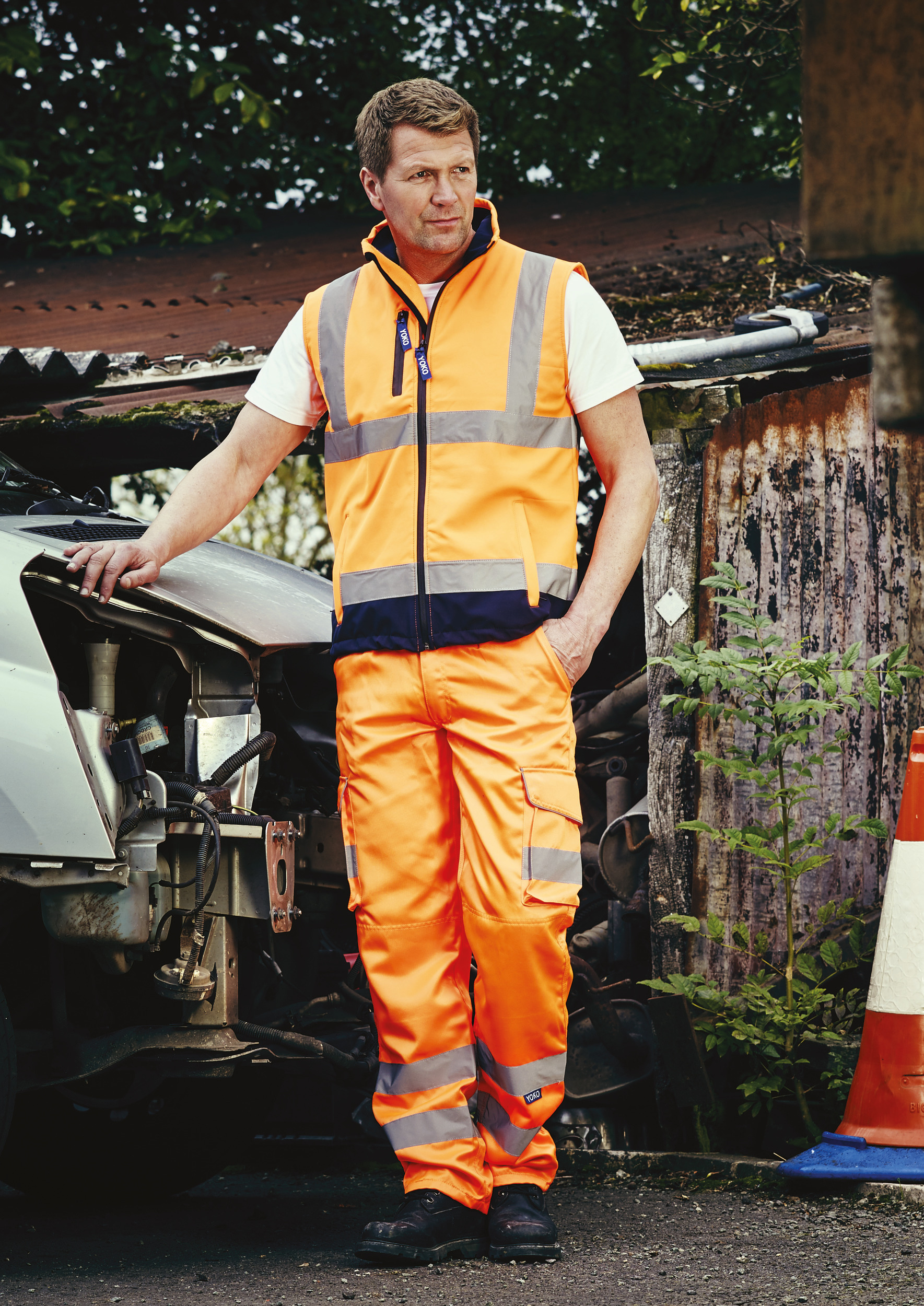 Hombre con chaleco y pantalón reflectante naranja apoyado en un vehículo averiado junto a un cono de tráfico en un entorno industrial.