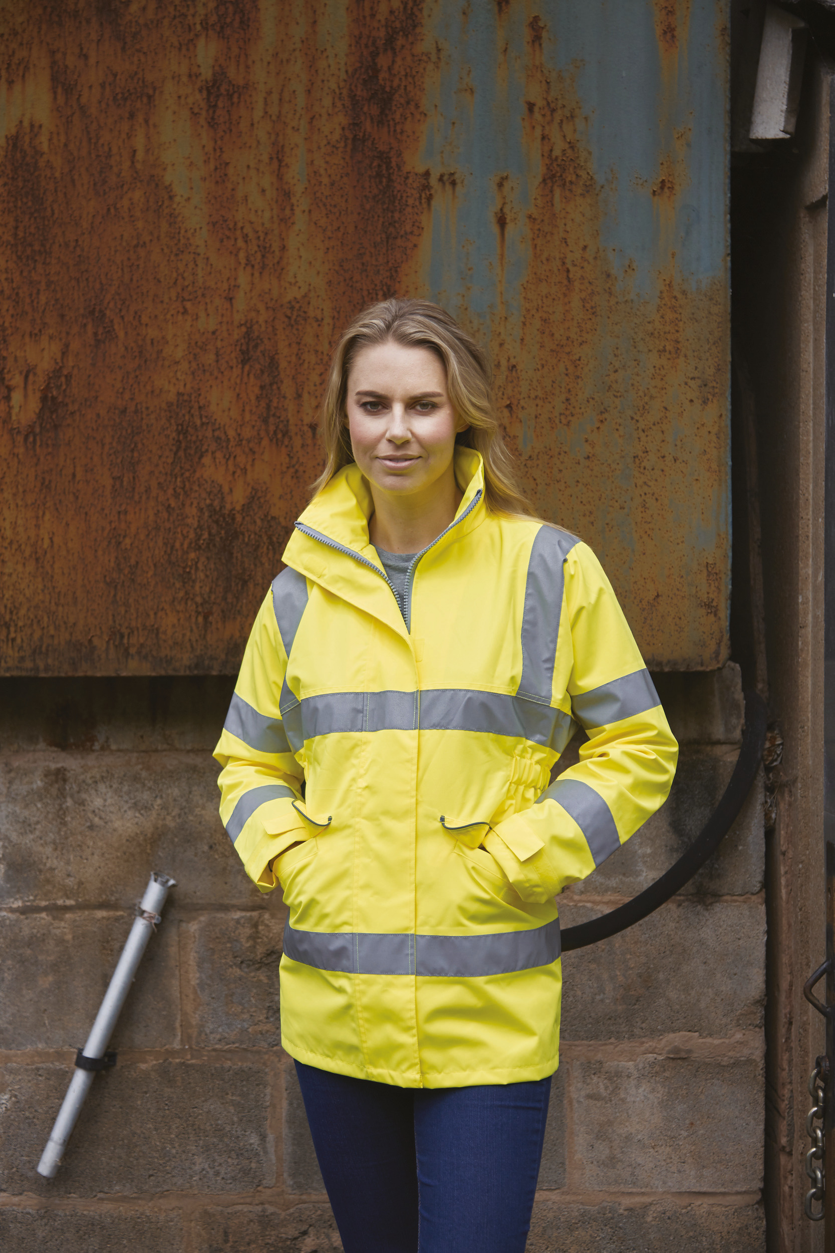 Mujer con chaqueta amarilla reflectante frente a una pared metálica oxidada en un entorno industrial