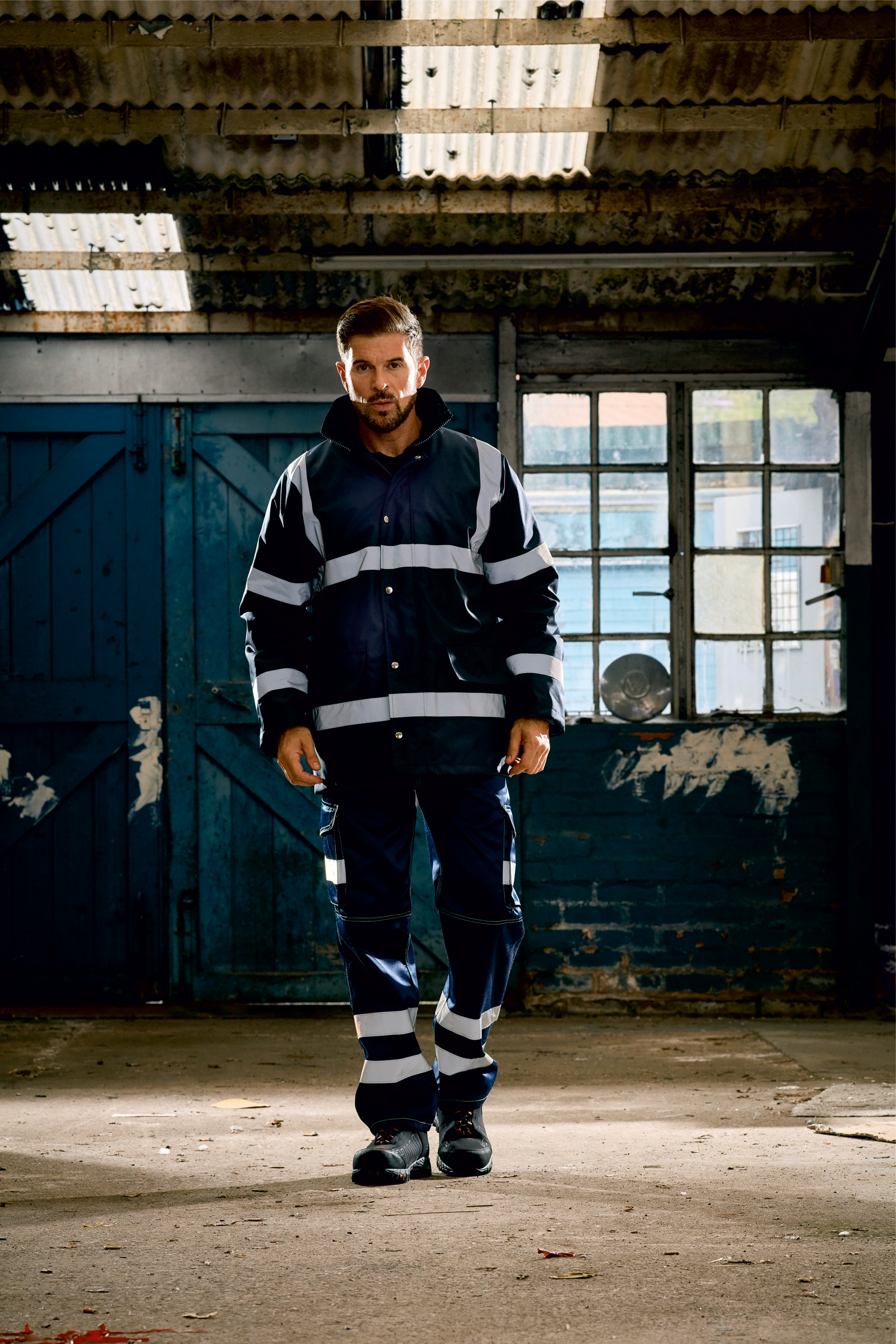 Hombre con chaqueta y pantalón de trabajo reflectantes en el interior de un taller industrial desvencijado