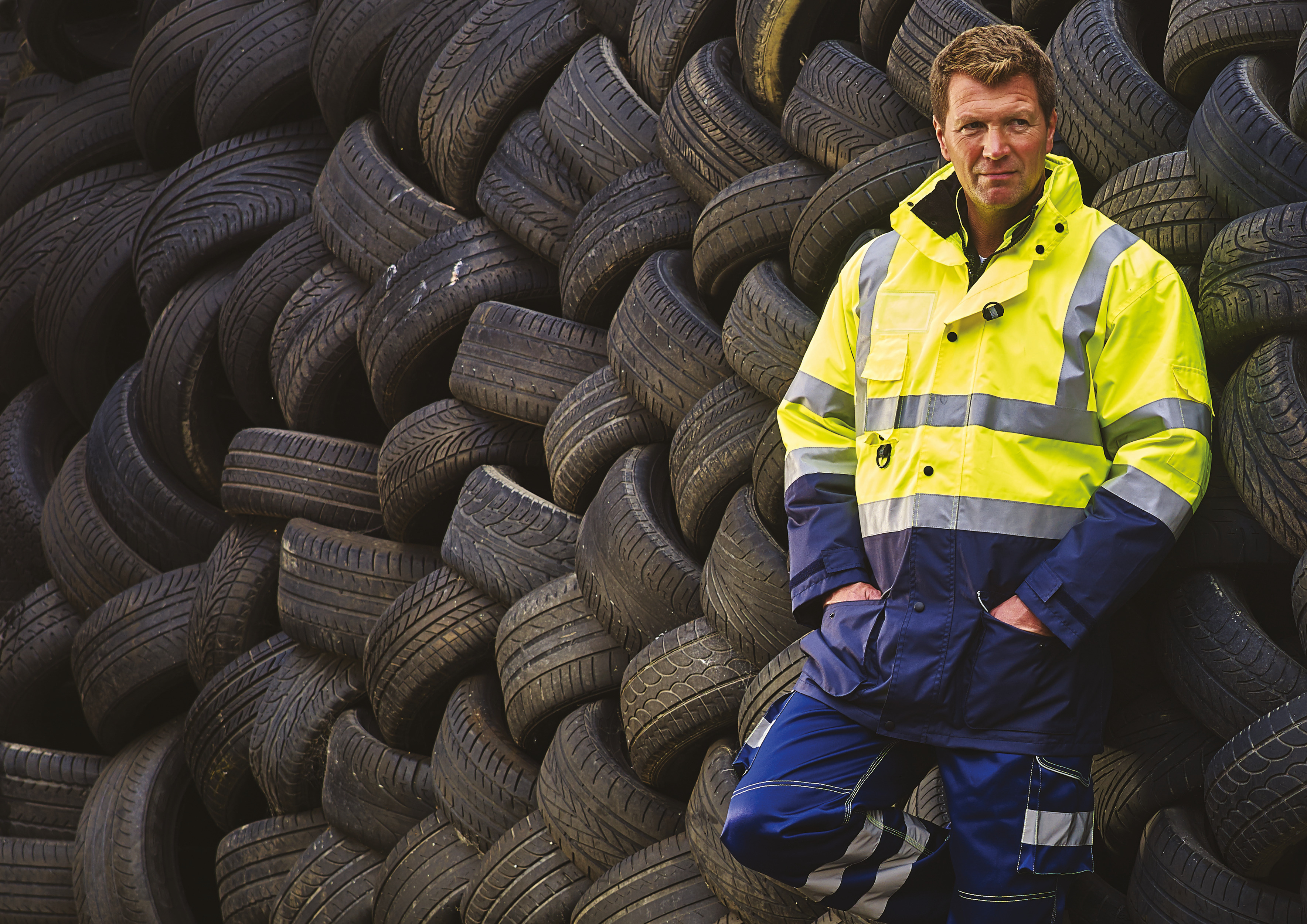 Hombre con chaqueta de alta visibilidad apoyado en una pila de neumáticos usados.