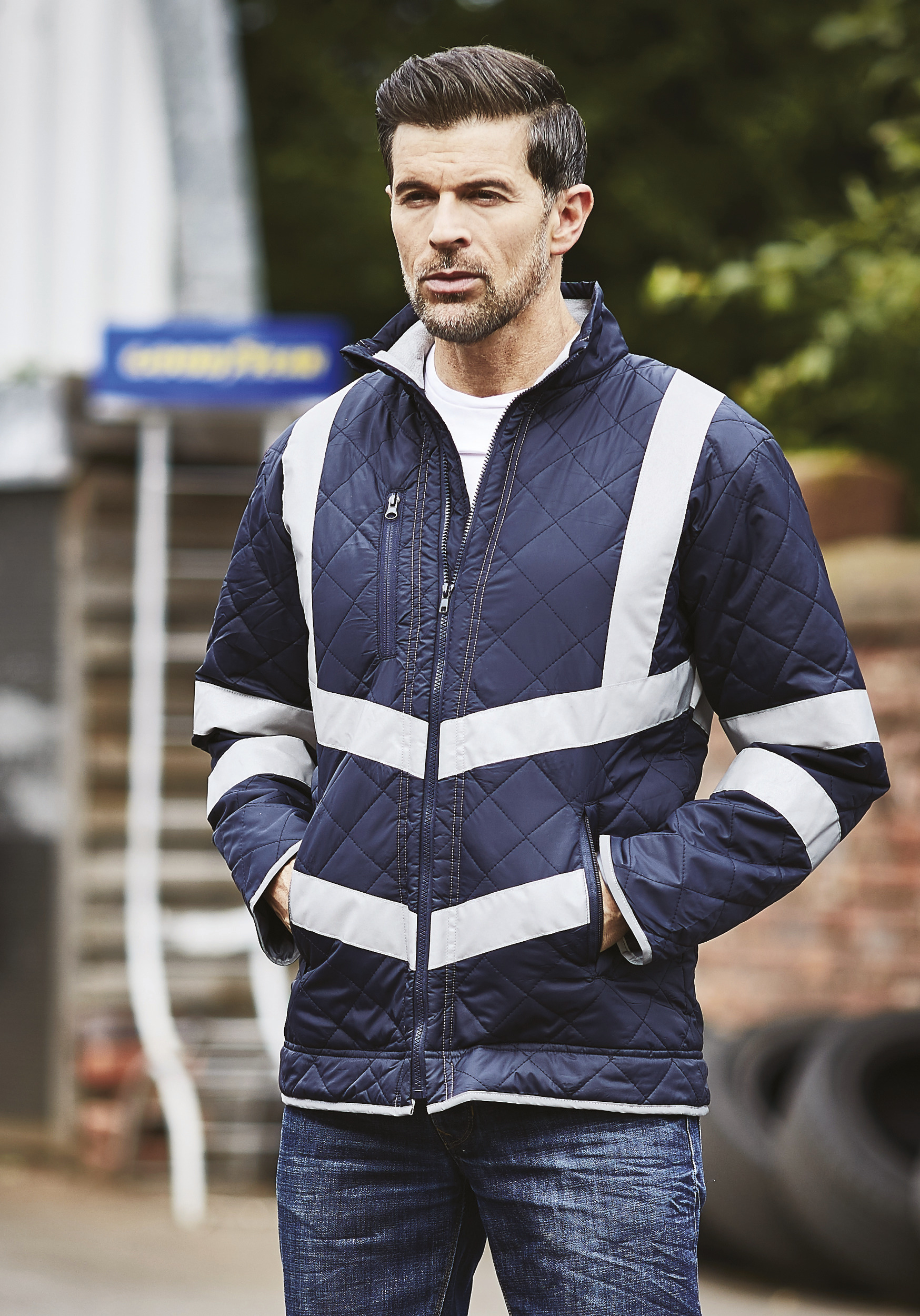 Hombre con chaqueta acolchada azul marino y franjas reflectantes de alta visibilidad en un entorno industrial.