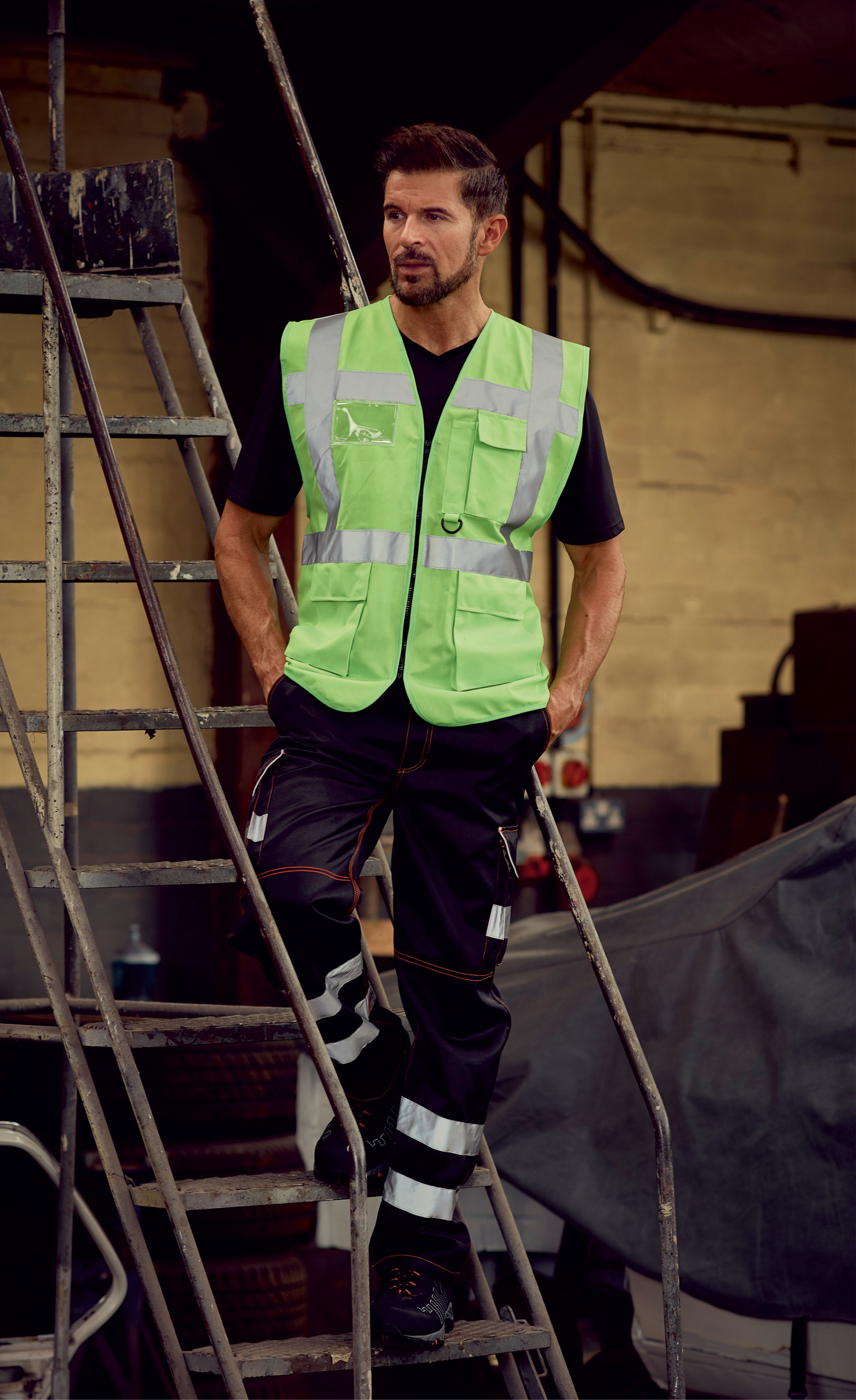 Hombre con chaleco reflectante verde y pantalones de trabajo subido en una escalera metálica en un taller industrial