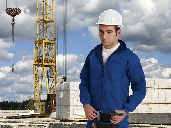 Obrero de la construcción con casco blanco y mono azul junto a grúa y materiales en obra al aire libre