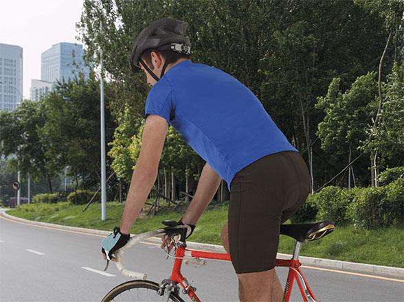 Ciclista con casco y camiseta azul pedaleando por una carretera urbana con edificios y árboles al fondo
