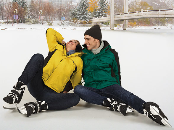 Pareja sentada sobre una pista de hielo al aire libre con chaquetas de invierno y paisaje nevado de fondo.