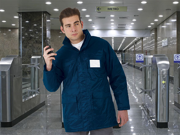 Empleado de metro con chaqueta azul junto a torniquetes hablando por walkie-talkie